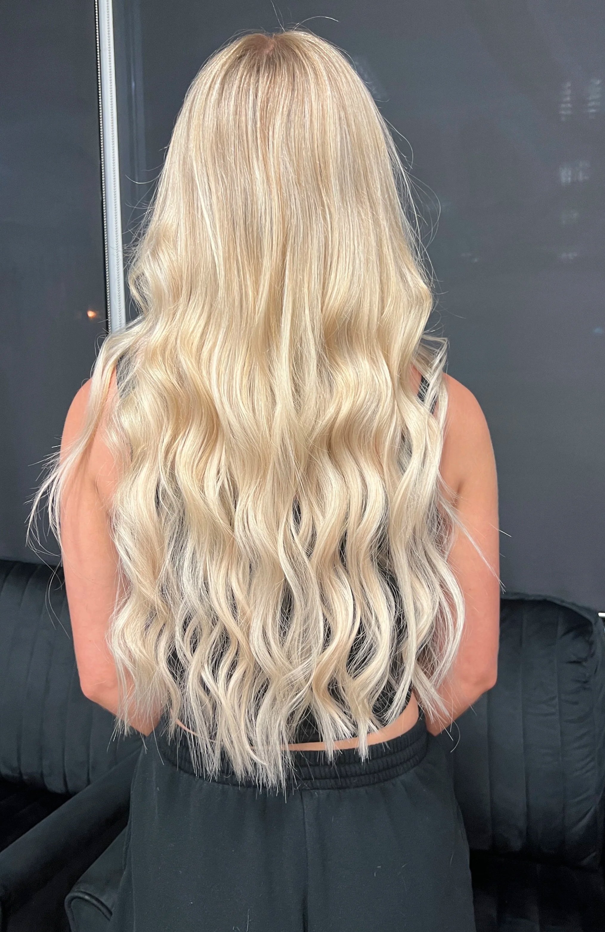 Back view of a woman with long, wavy blonde hair extensions, wearing a black top, sitting on a black sofa.
