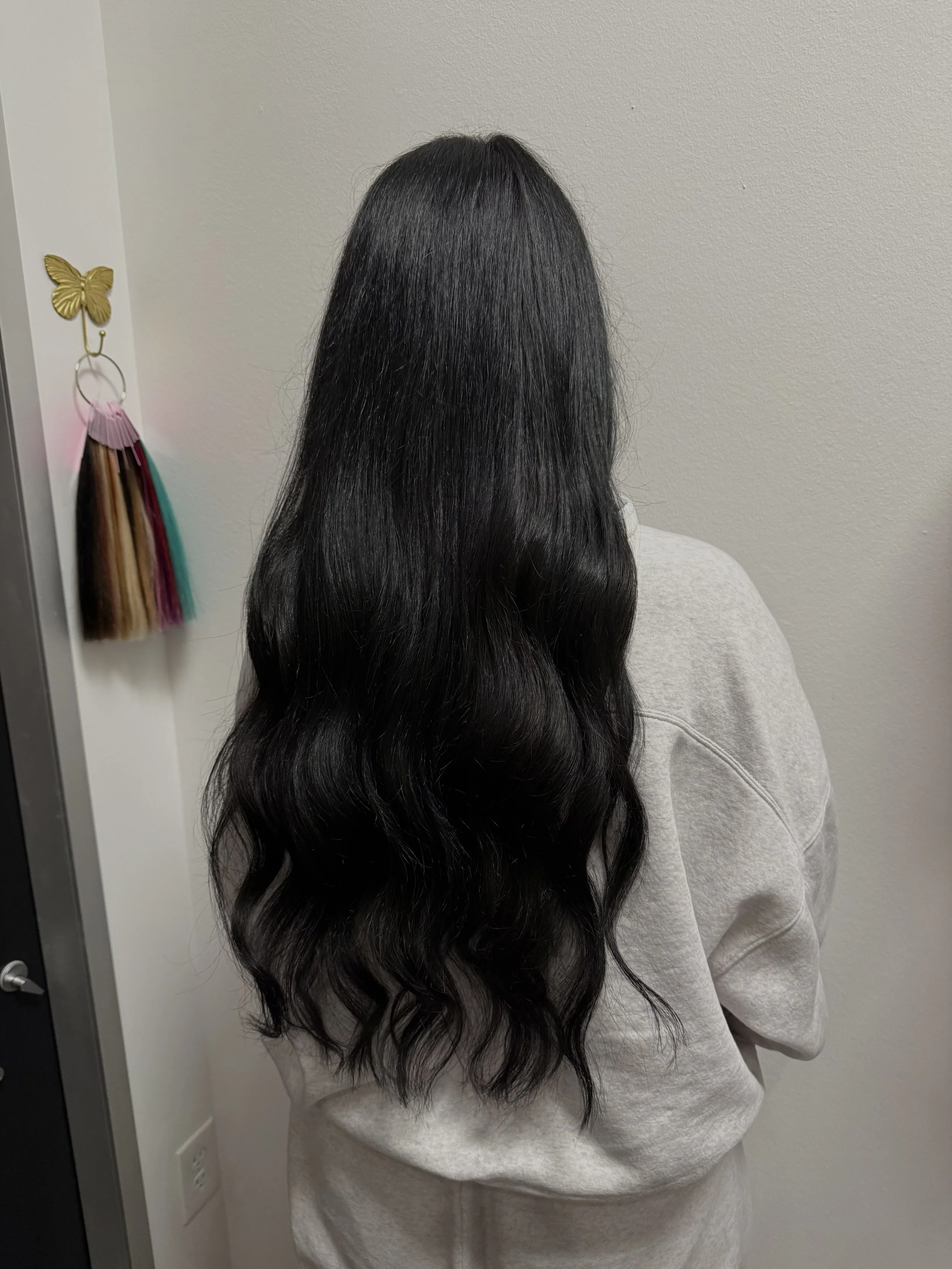 Back of a woman with long, black, wavy hair wearing a gray sweatshirt, standing in front of a white wall with a butterfly-shaped hook holding colored hair extensions.