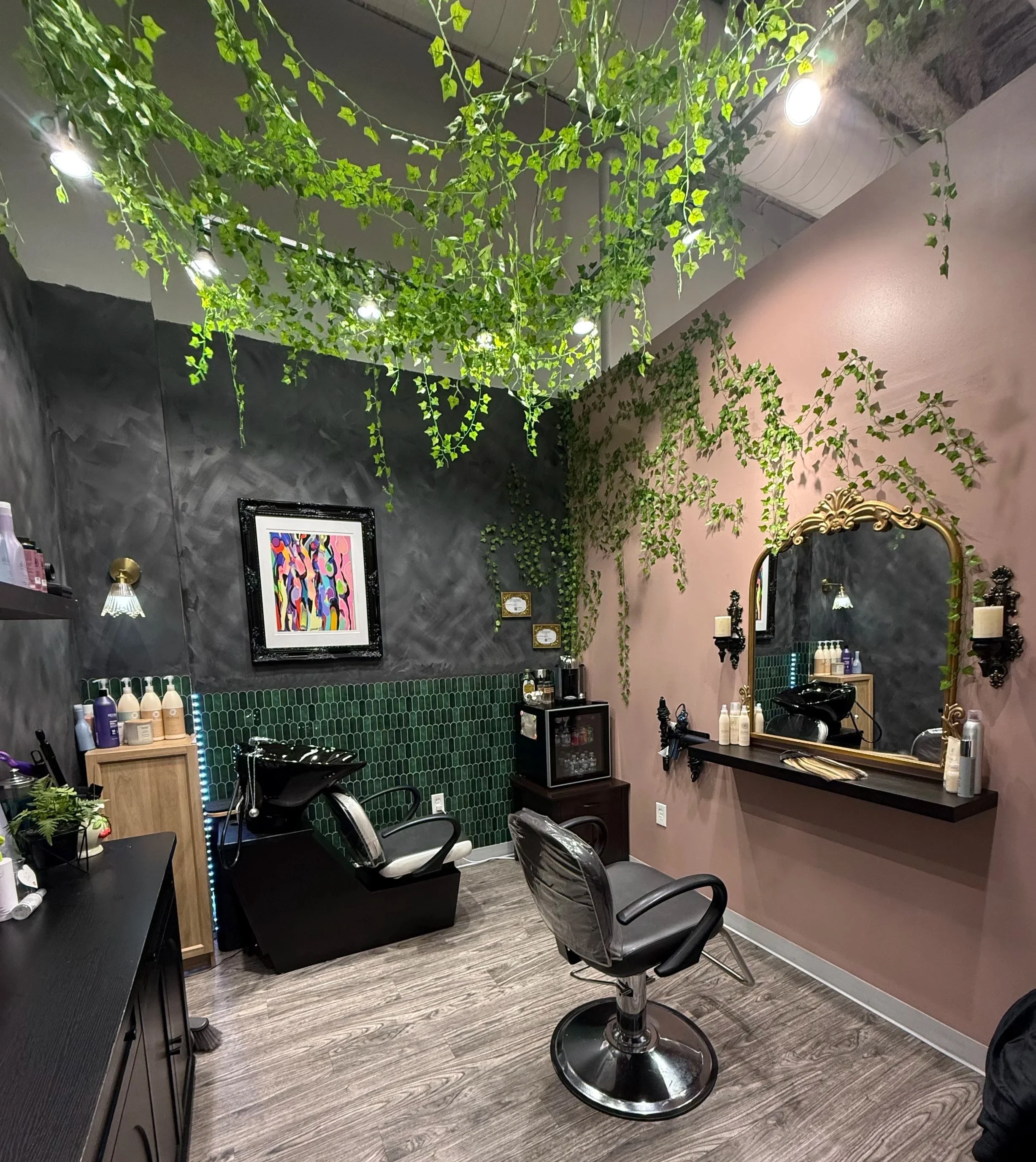 A modern salon interior with black and pink walls. The ceiling is decorated with hanging green ivy plants and spotlights. There are framed colorful abstract artworks on the wall, and a large mirror with a gold ornate frame. Speech bubbles are mounted on the wall with some text. There are salon chairs, a black counter, and various hair care products visible.
