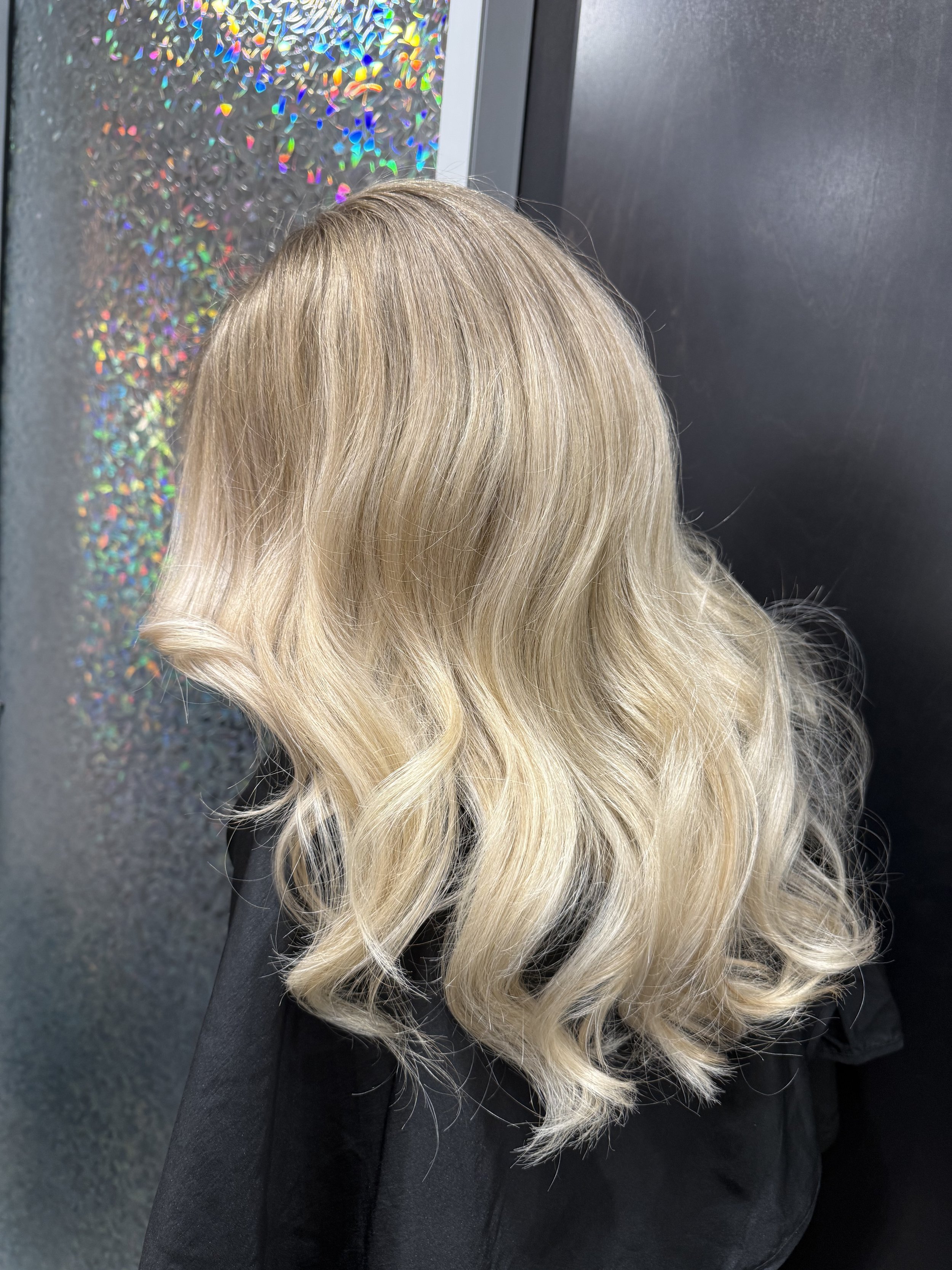 The image shows the back of a person's head with long, wavy blonde hair, wearing a black jacket, standing in front of a textured black wall and a reflective, iridescent panel.