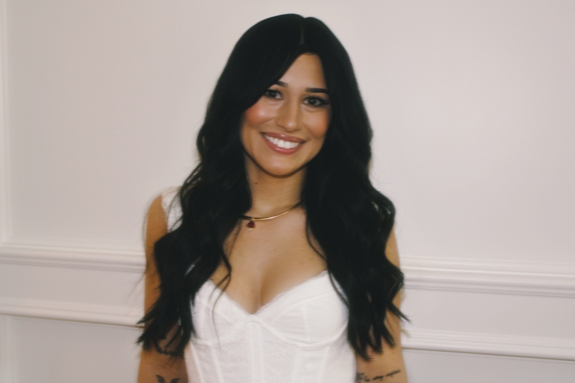 A young woman with long black hair, smiling, wearing a white top and a gold necklace, standing against a plain light-colored wall.