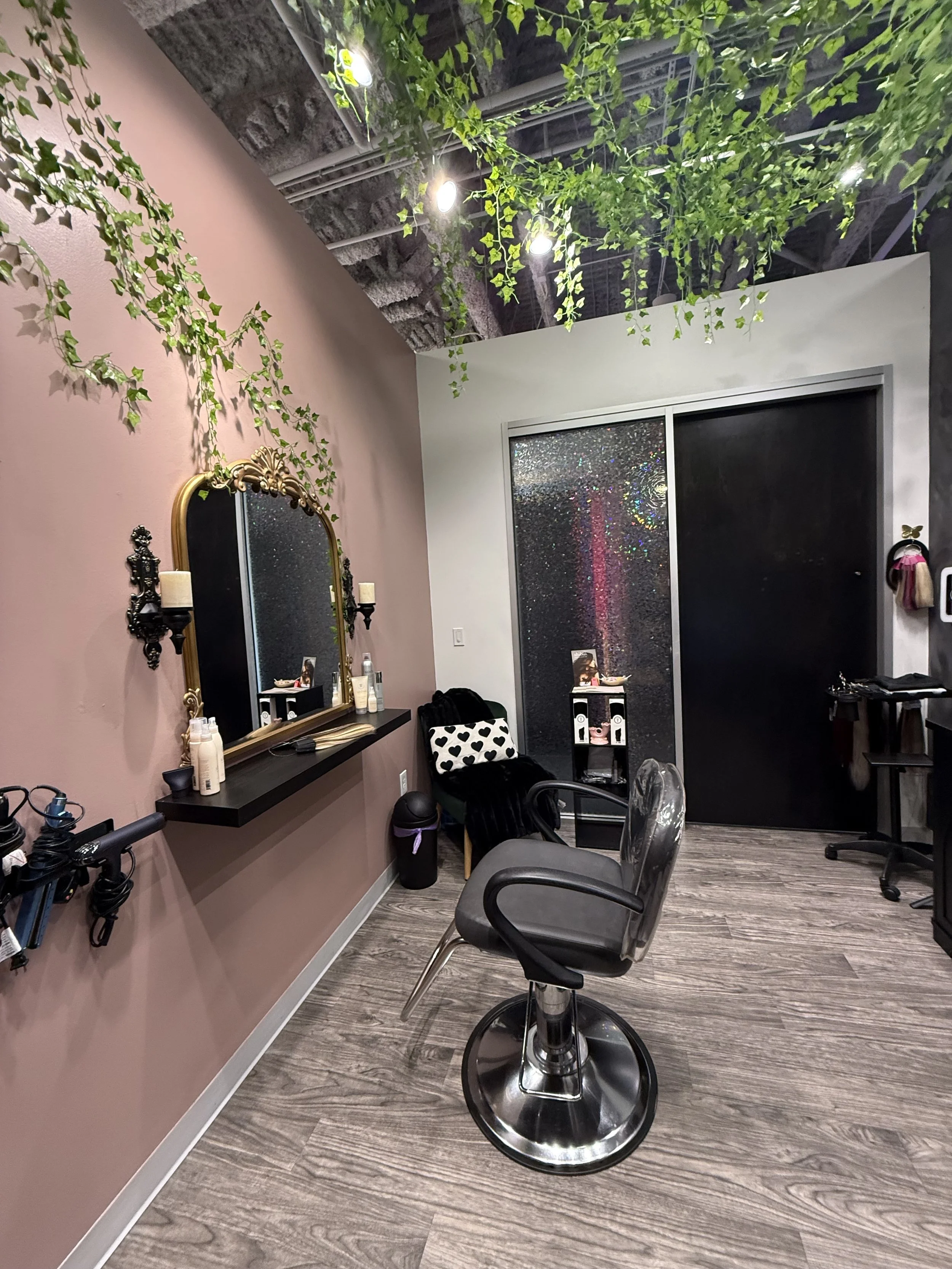 A salon or spa interior with a styling chair, large mirror with gold trim, and green leafy decorations hanging from the ceiling. The room has a pink and white wall, and dark wood flooring. There is a black shelf with beauty products and a black chair with polka dot cushion. The back wall has a sliding glass door with rain droplets.