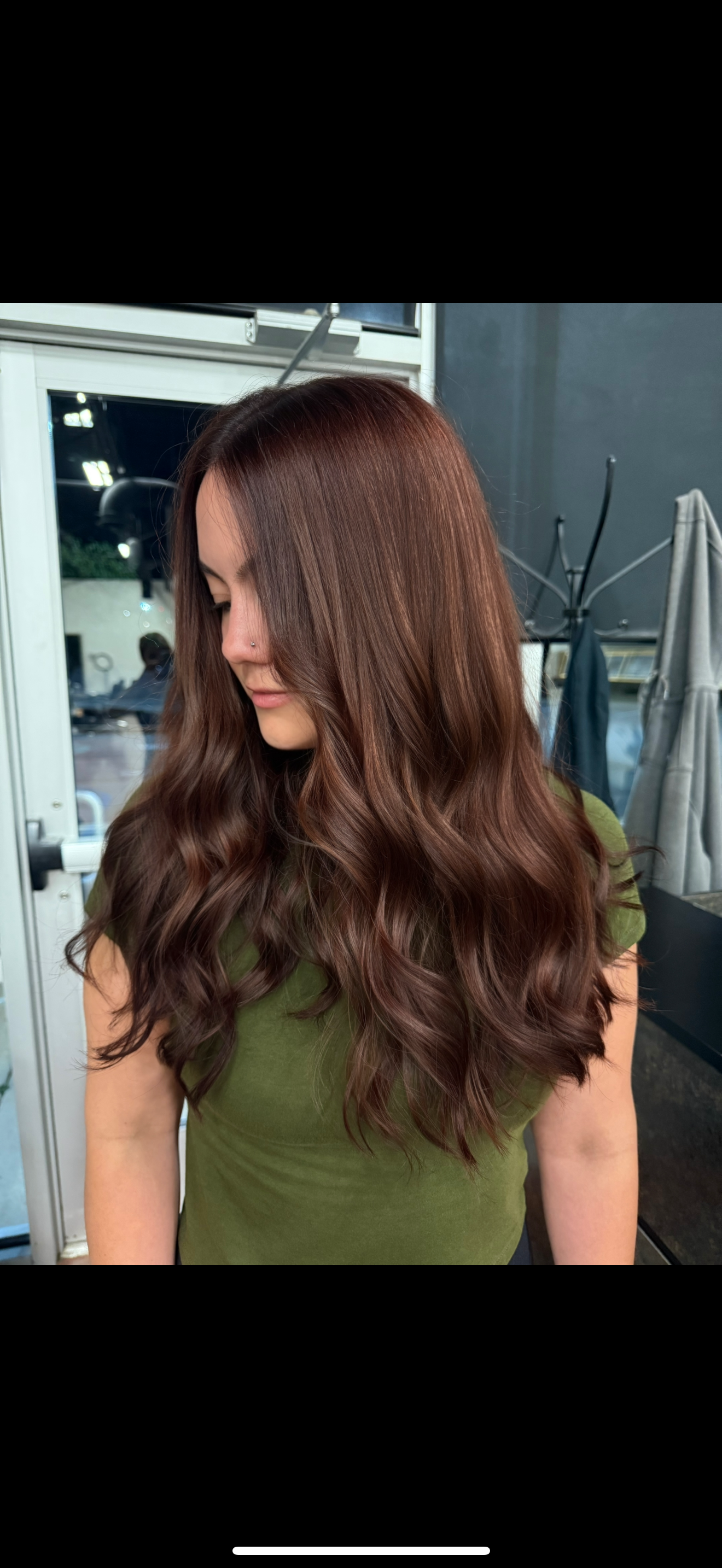 A woman with long, wavy, reddish-brown hair looking down and away, wearing an olive green shirt, indoors with a window and coat rack in the background.