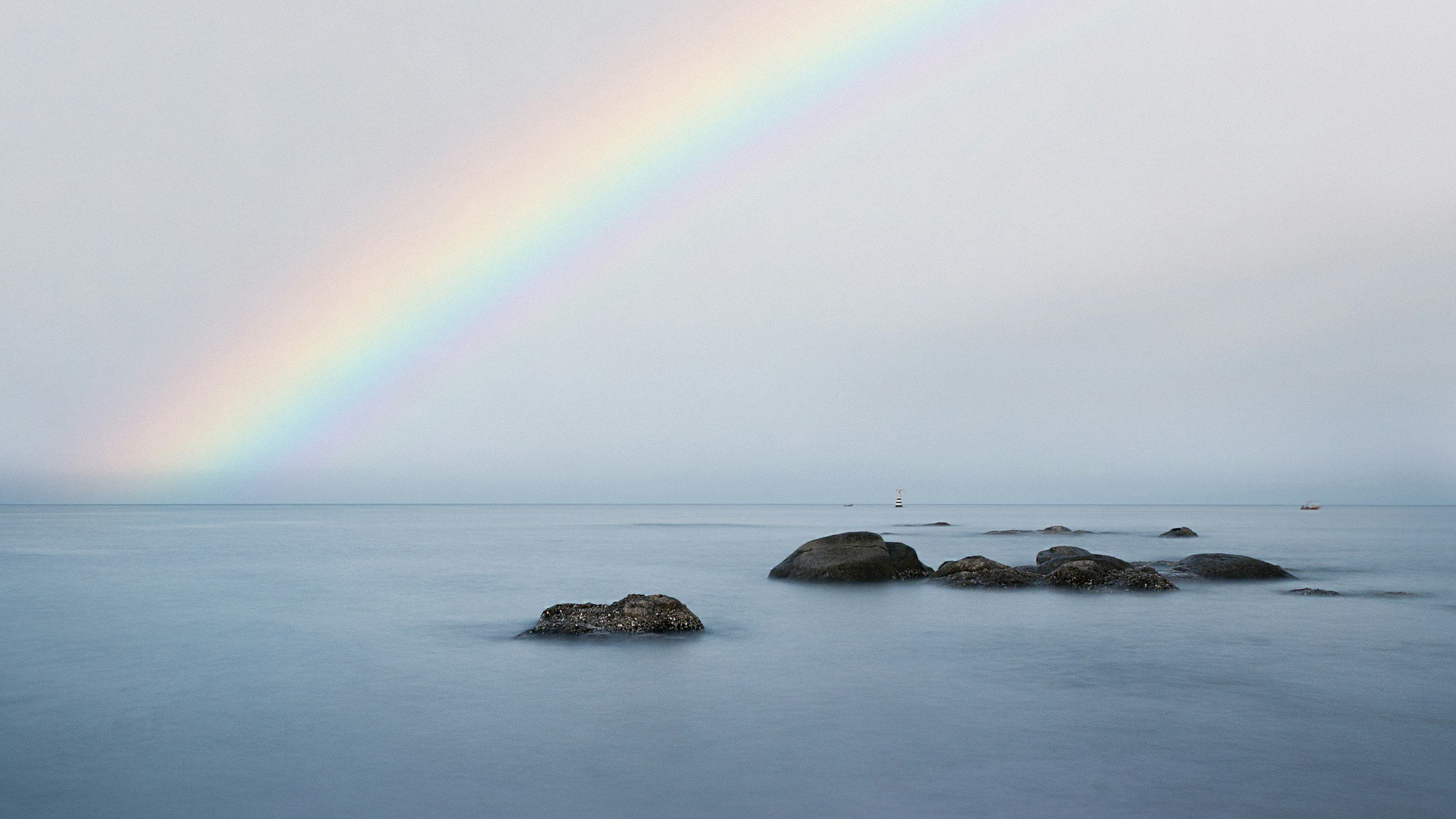 Calm ocean with multiple large rocks near the shore, a lighthouse in the distance, and a rainbow arching across a gray sky.