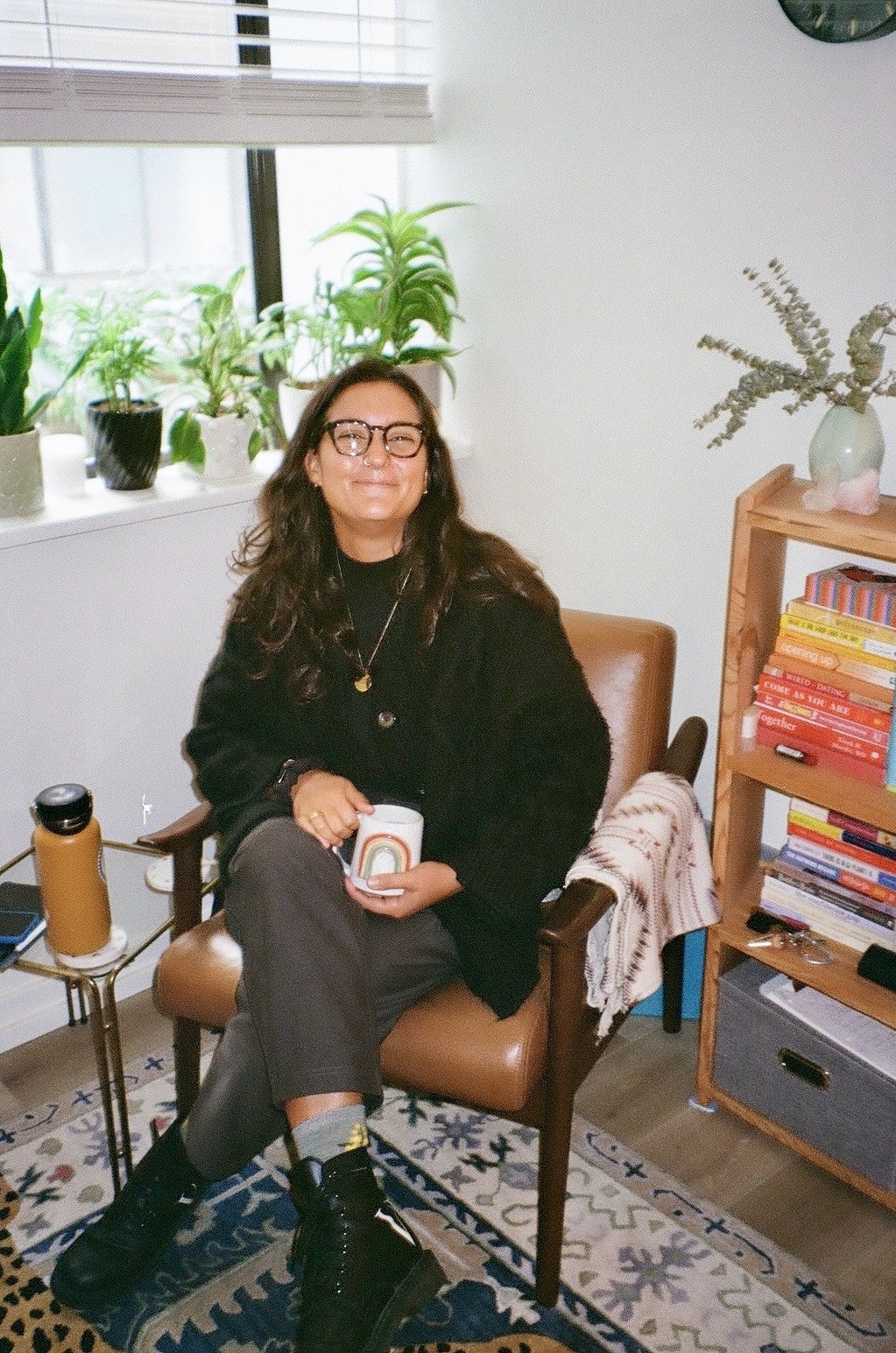 Woman sitting in a brown chair holding a mug, smiling, in a room with houseplants on the window sill, books on a wooden shelf, and a patterned rug on the floor.