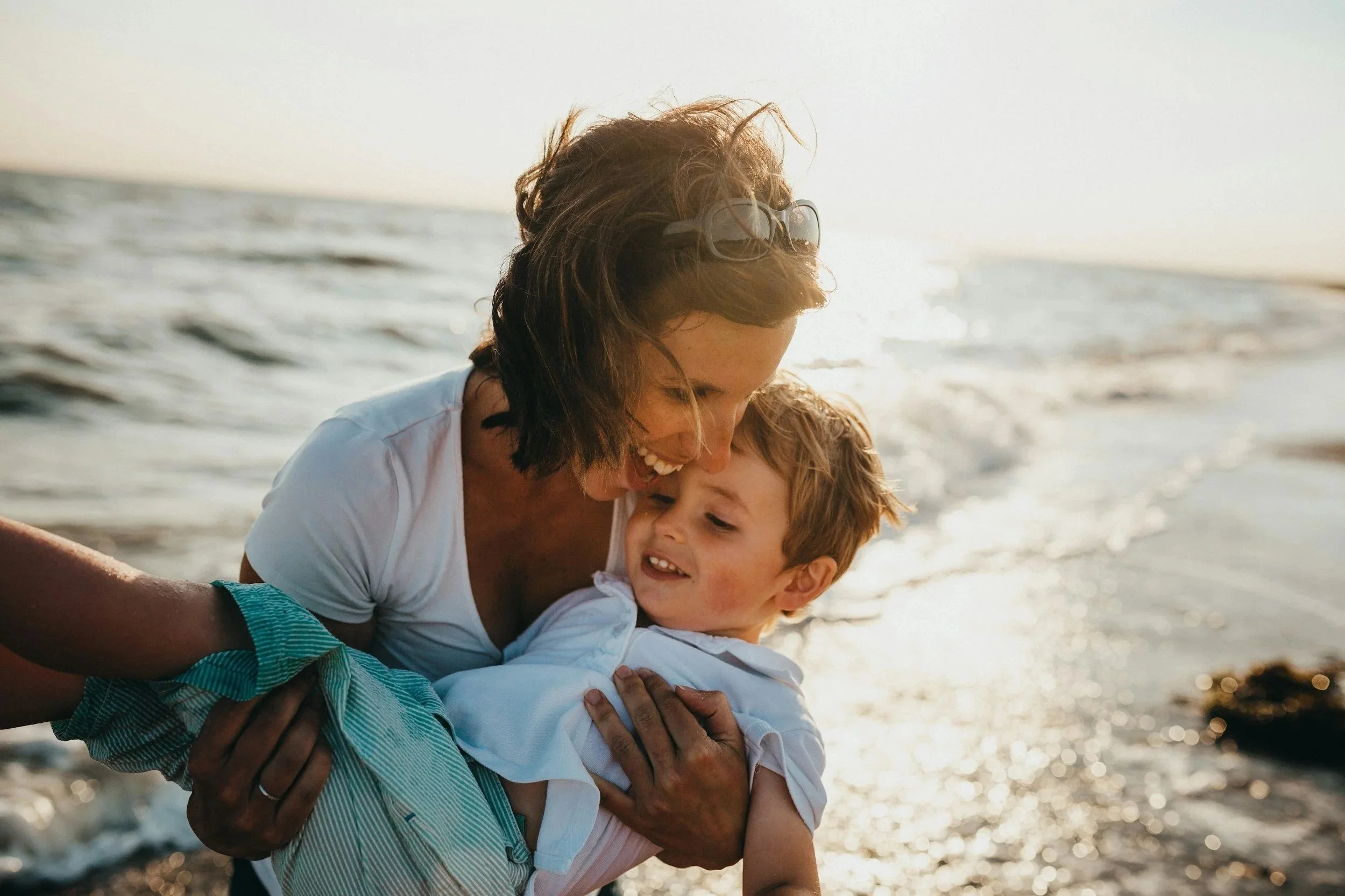 A woman and a young boy joyfully embrace by the beach during sunset, with ocean waves in the background.