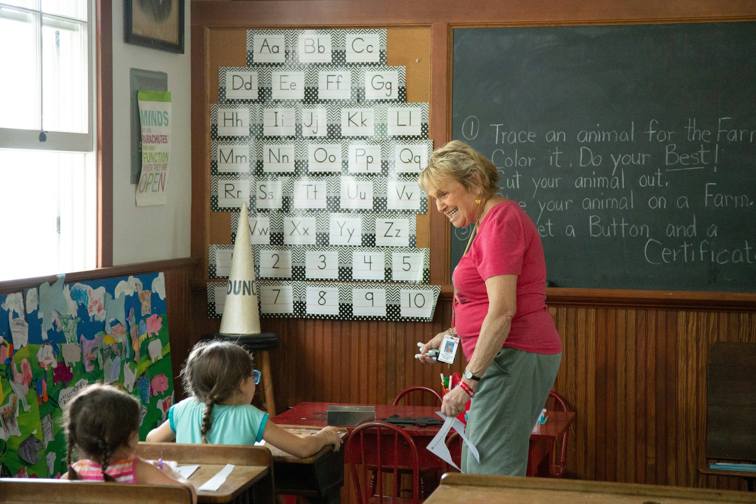 A classroom with children sitting at desks and a teacher standing near a chalkboard. There is an alphabet display on the wall behind the teacher, showing letters A through Z with decorative backgrounds. The teacher is smiling and engaging with the students.