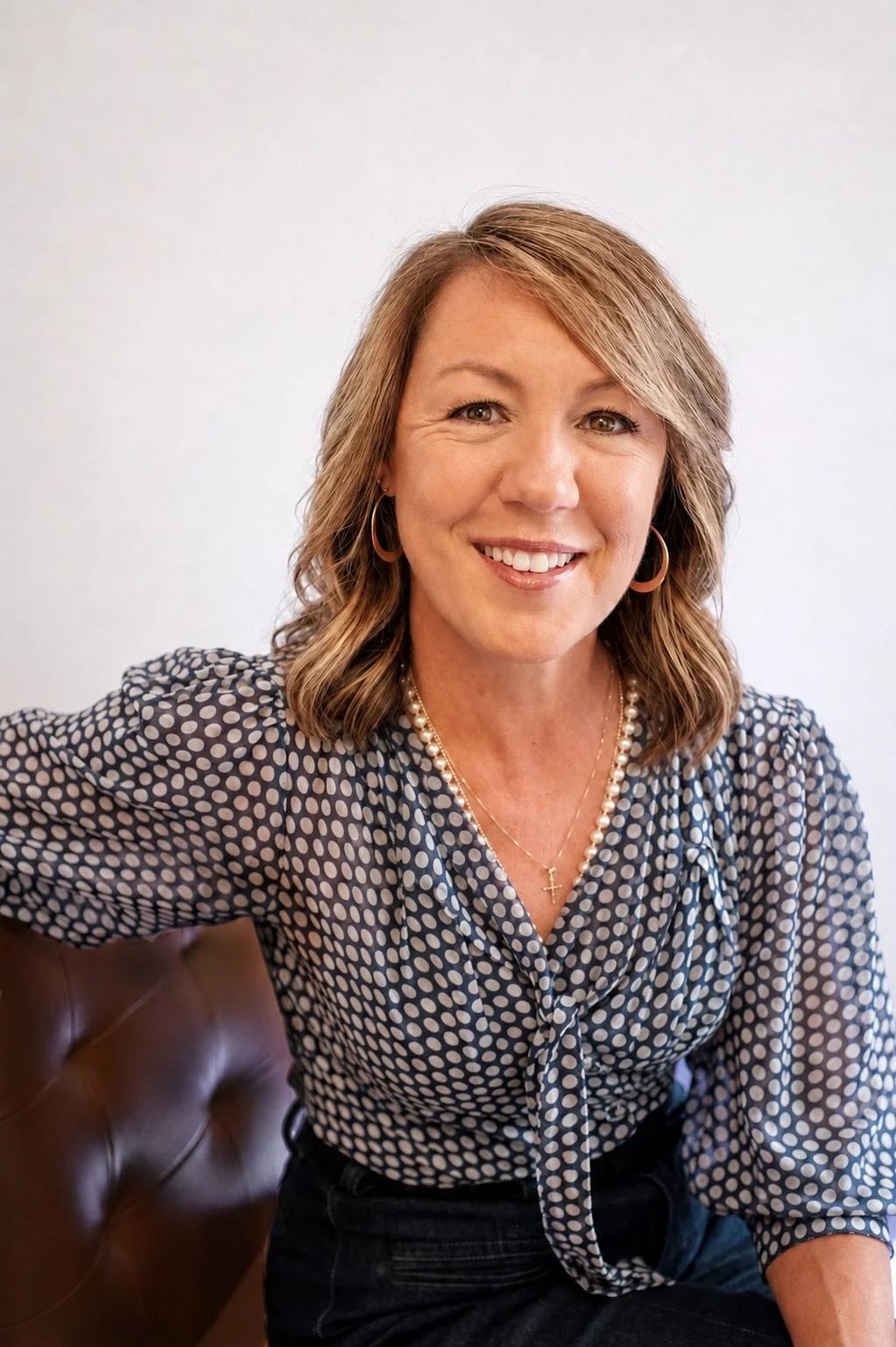 A woman with shoulder-length blonde hair smiling, wearing hoop earrings, a pearl necklace, and a polka-dot blouse.