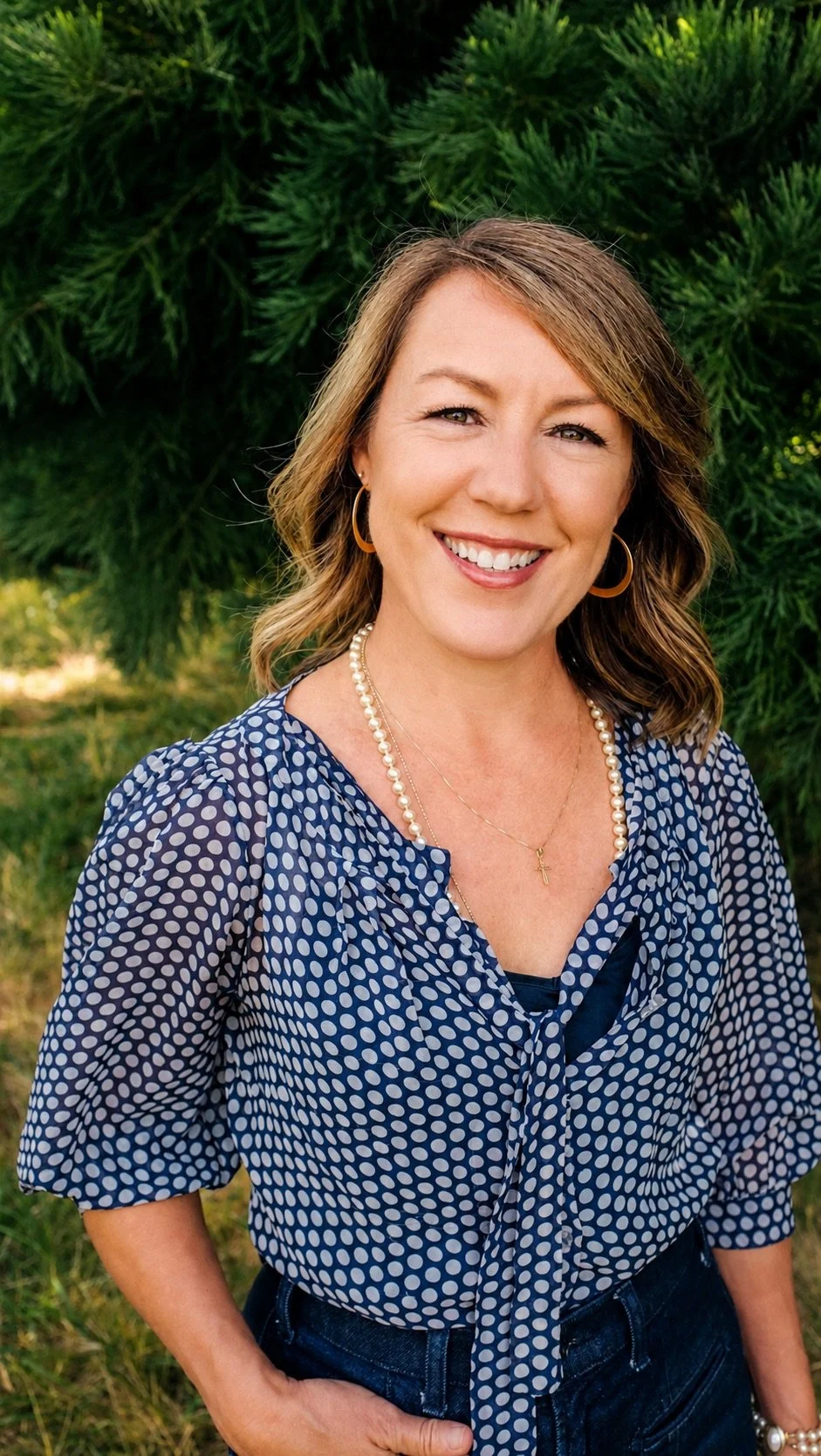 A woman with shoulder-length wavy hair wearing a blue polka dot blouse, pearl necklace, gold cross necklace, gold hoop earrings, and smiling outdoors in front of green trees.
