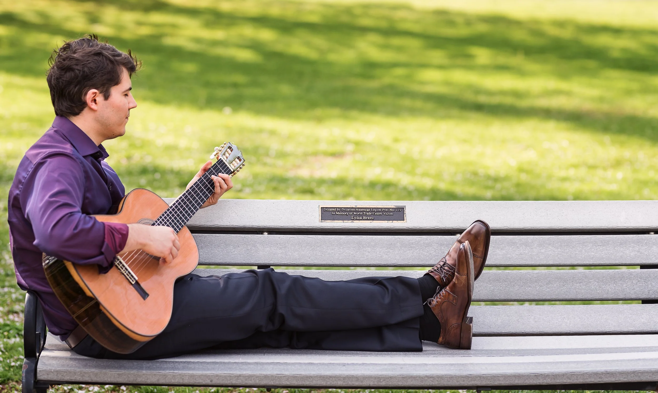 A man in a purple shirt and black pants lounges on a park bench, playing an acoustic guitar. His legs are extended on the bench, wearing brown leather shoes. A plaque on the bench commemorates Lydia Bravo. The background features a green, leafy park.