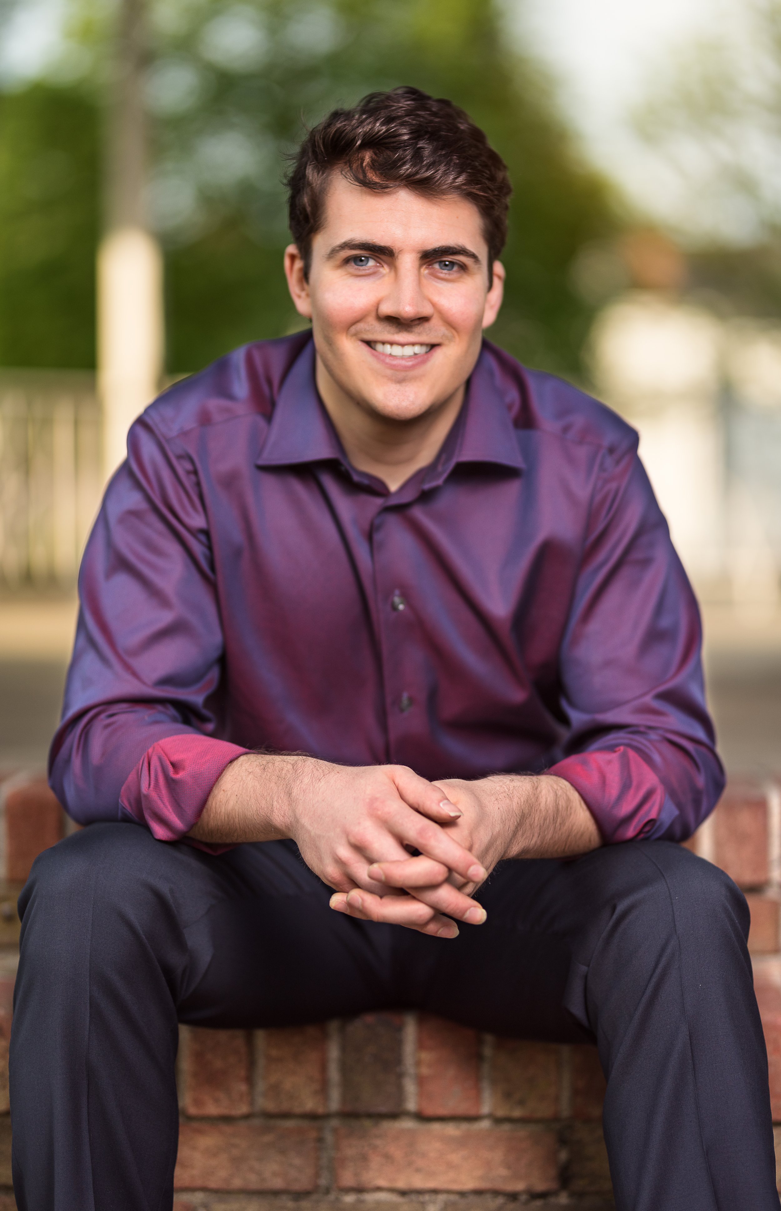 Smiling young man in a purple dress shirt sitting on a brick step outdoors, with greenery in the background.