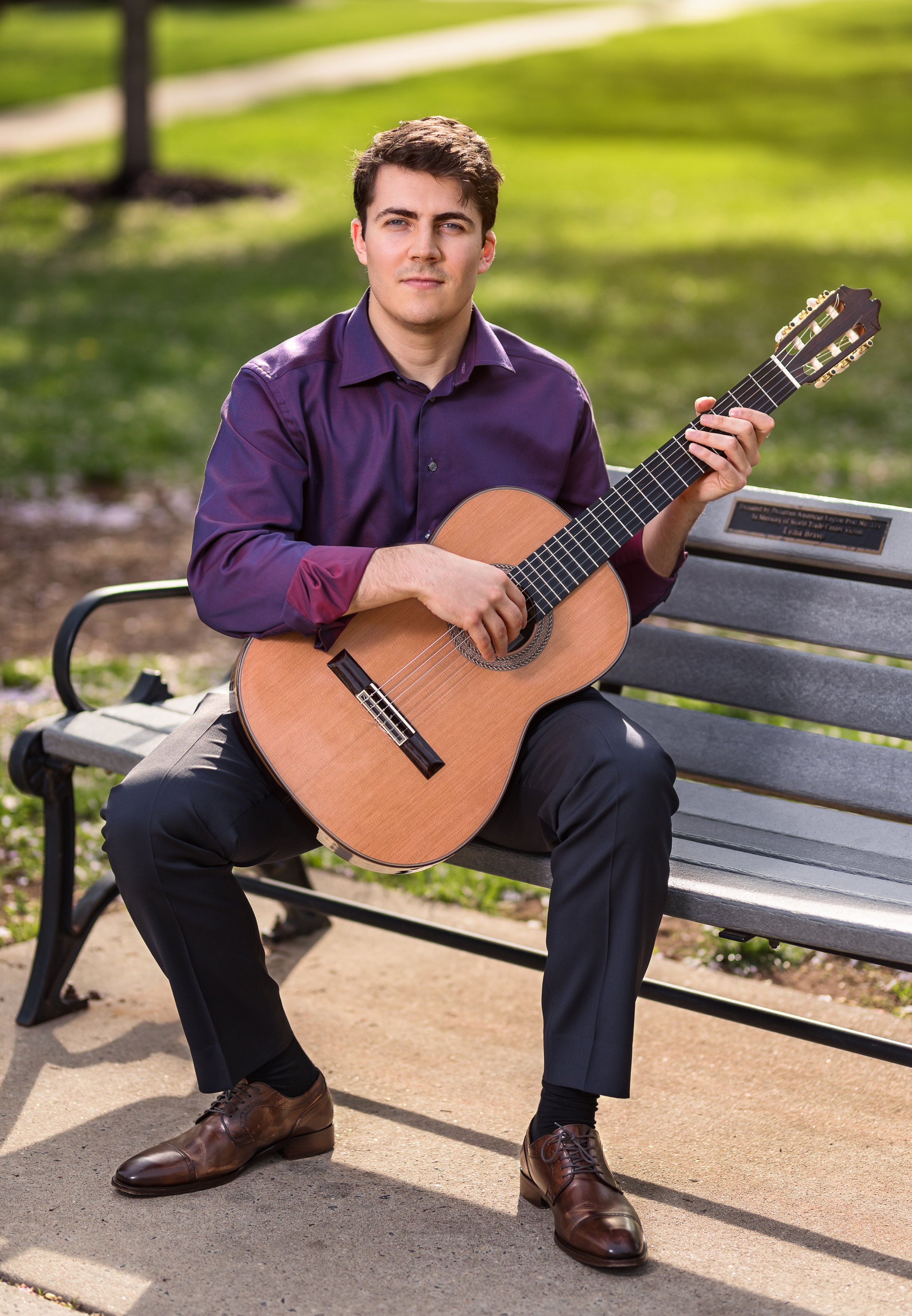 A young man sitting on a park bench playing an acoustic guitar, wearing a purple shirt and black pants, with greenery in the background.