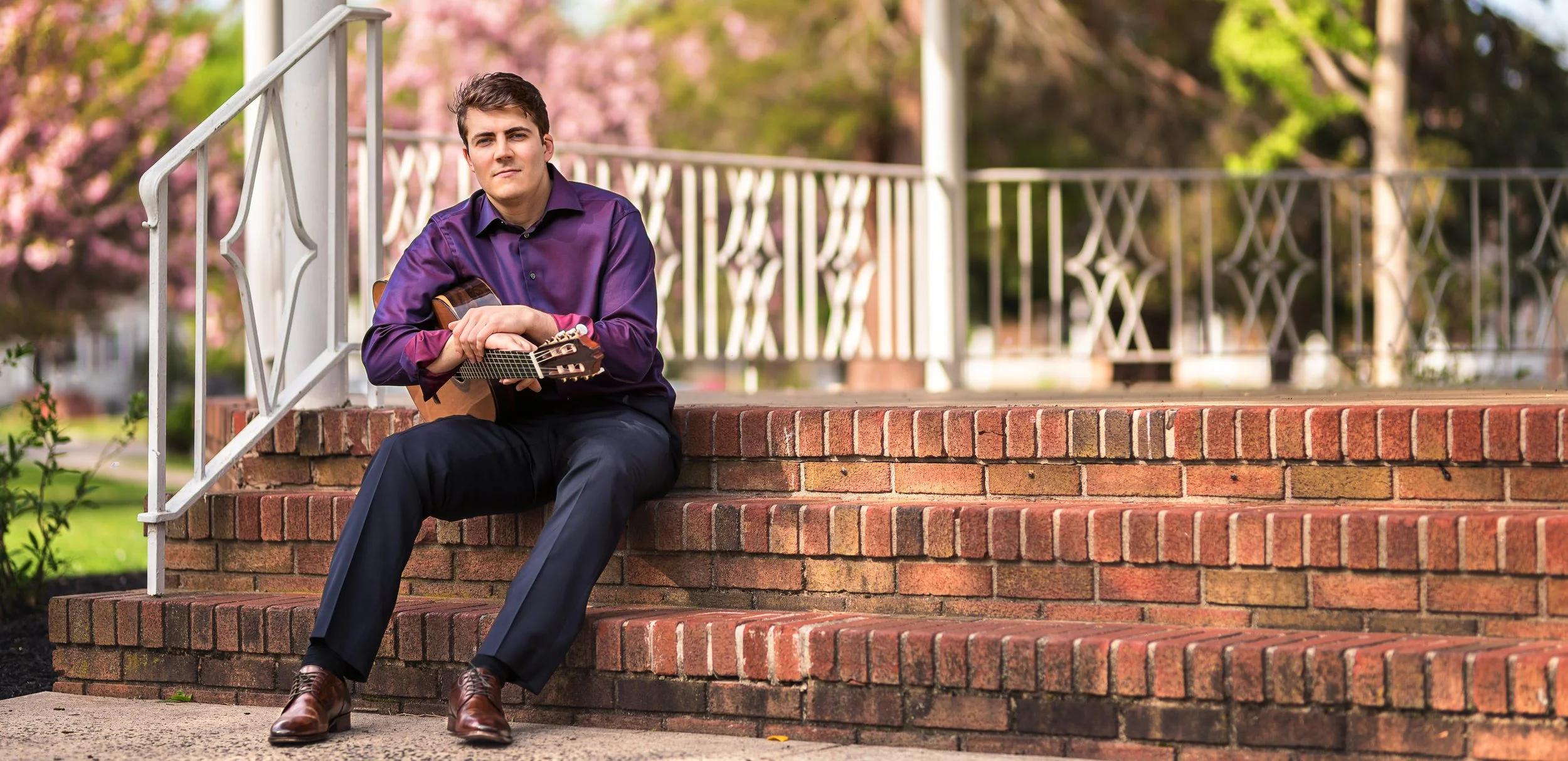 A young man in a purple shirt and dark pants sitting on brick steps outdoors, holding an acoustic guitar, with trees and pink blossoms in the background.