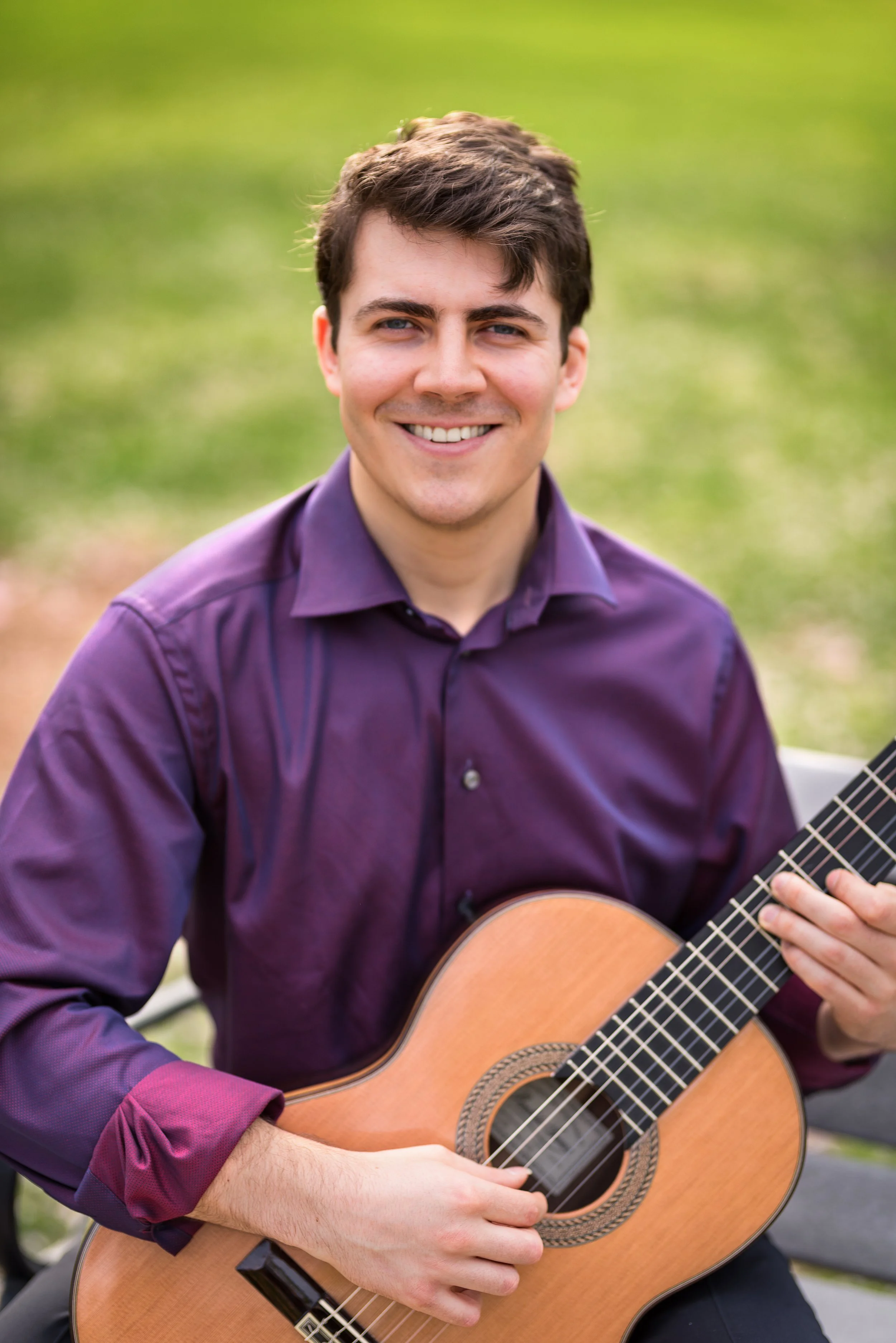 Young man playing an acoustic guitar outdoors, wearing a purple shirt, smiling, with green blurred background.