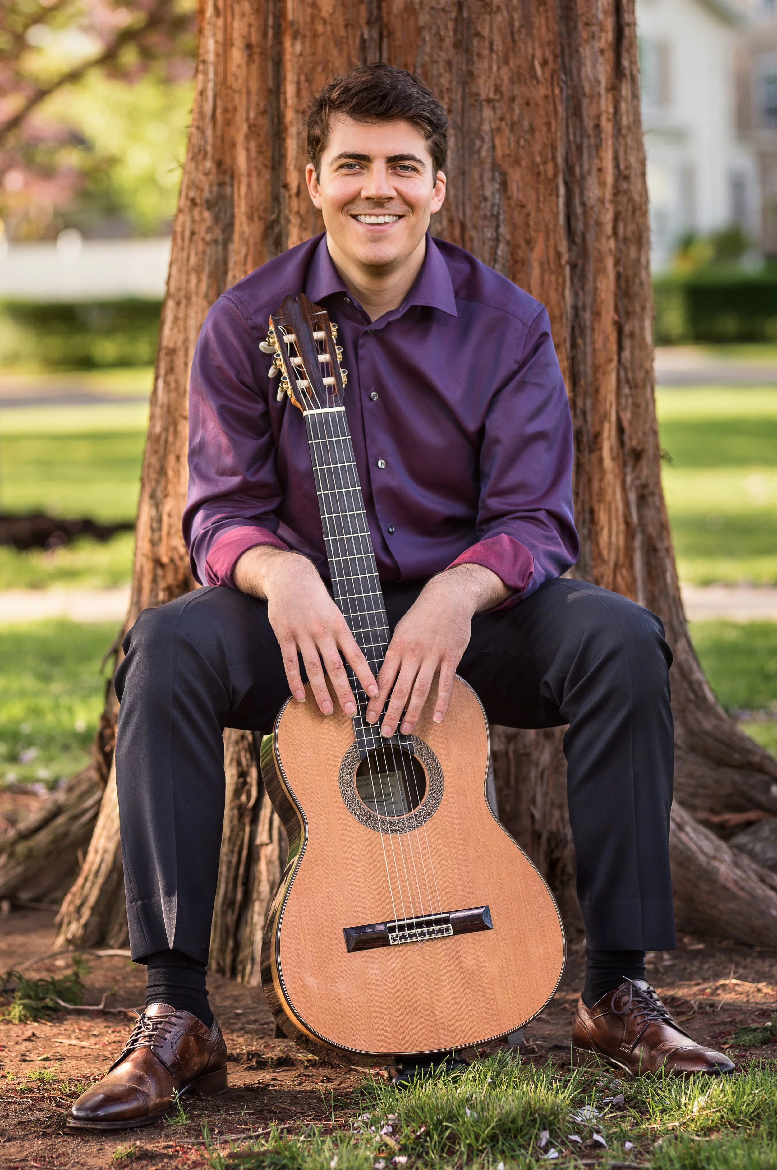 A young man with dark hair, wearing a purple shirt and black pants, sitting outdoors in front of a large tree. He is smiling and holding an acoustic guitar, with the guitar resting on the ground between his legs.
