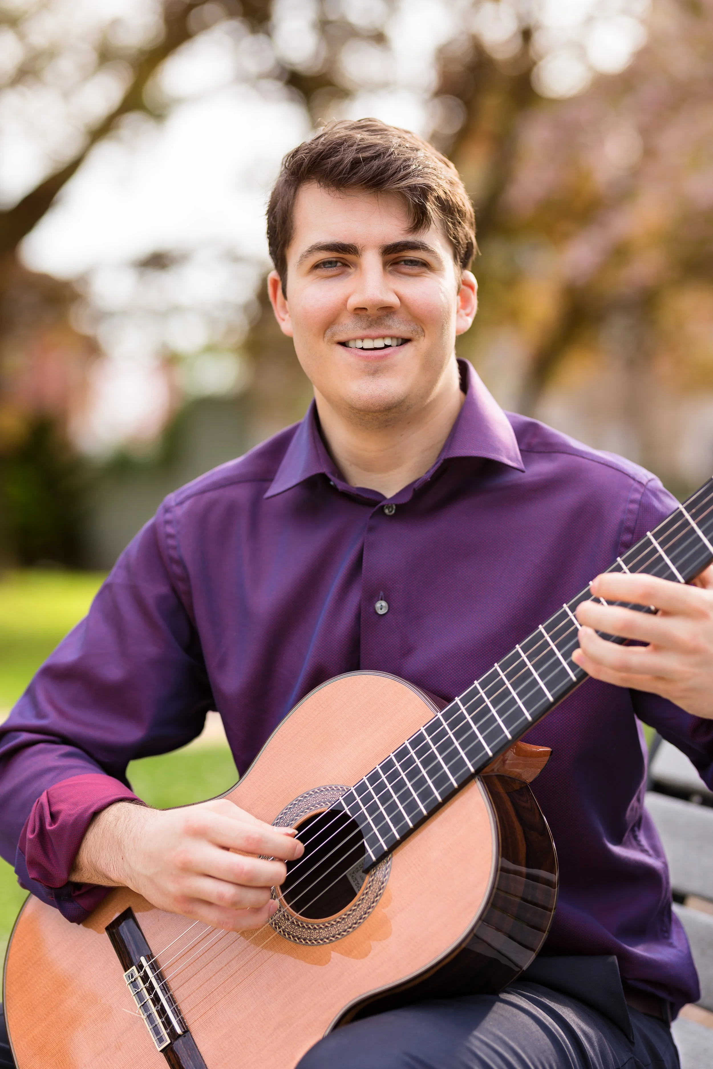 A young man with brown hair wearing a purple shirt, playing an acoustic guitar outdoors in a park with trees and pink blossoms in the background.
