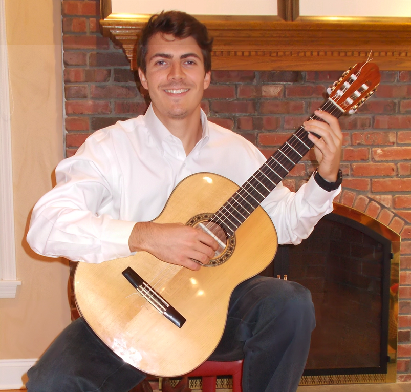 A man smiling and playing an acoustic guitar indoors in front of a brick fireplace.