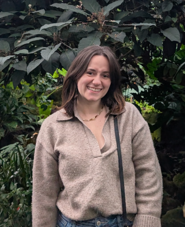 A woman with shoulder-length brown hair smiling, wearing a beige sweater and a necklace, standing outdoors in front of large green leaves.