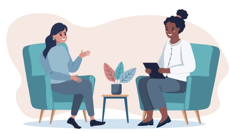 Two women having a conversation seated in teal armchairs with a small table and plant between them.