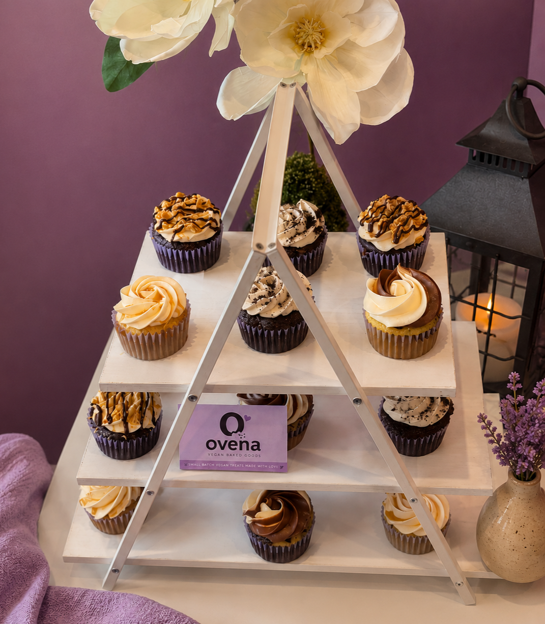 A white, three-tiered cupcake stand holds ten cupcakes with swirled frosting and chocolate drizzle, decorated with caramel, cookies, and chocolate. The stand is near a purple wall, with large white flowers and a black lantern with a candle, along with a purple flower arrangement in a beige vase.