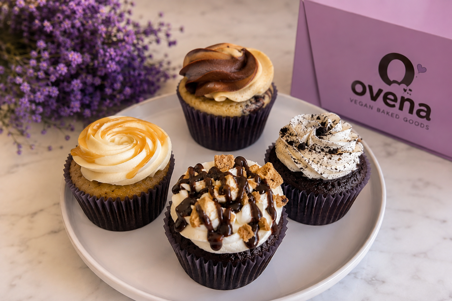 Four vegan cupcakes on a white plate with purple flowering plant and a purple box in the background.