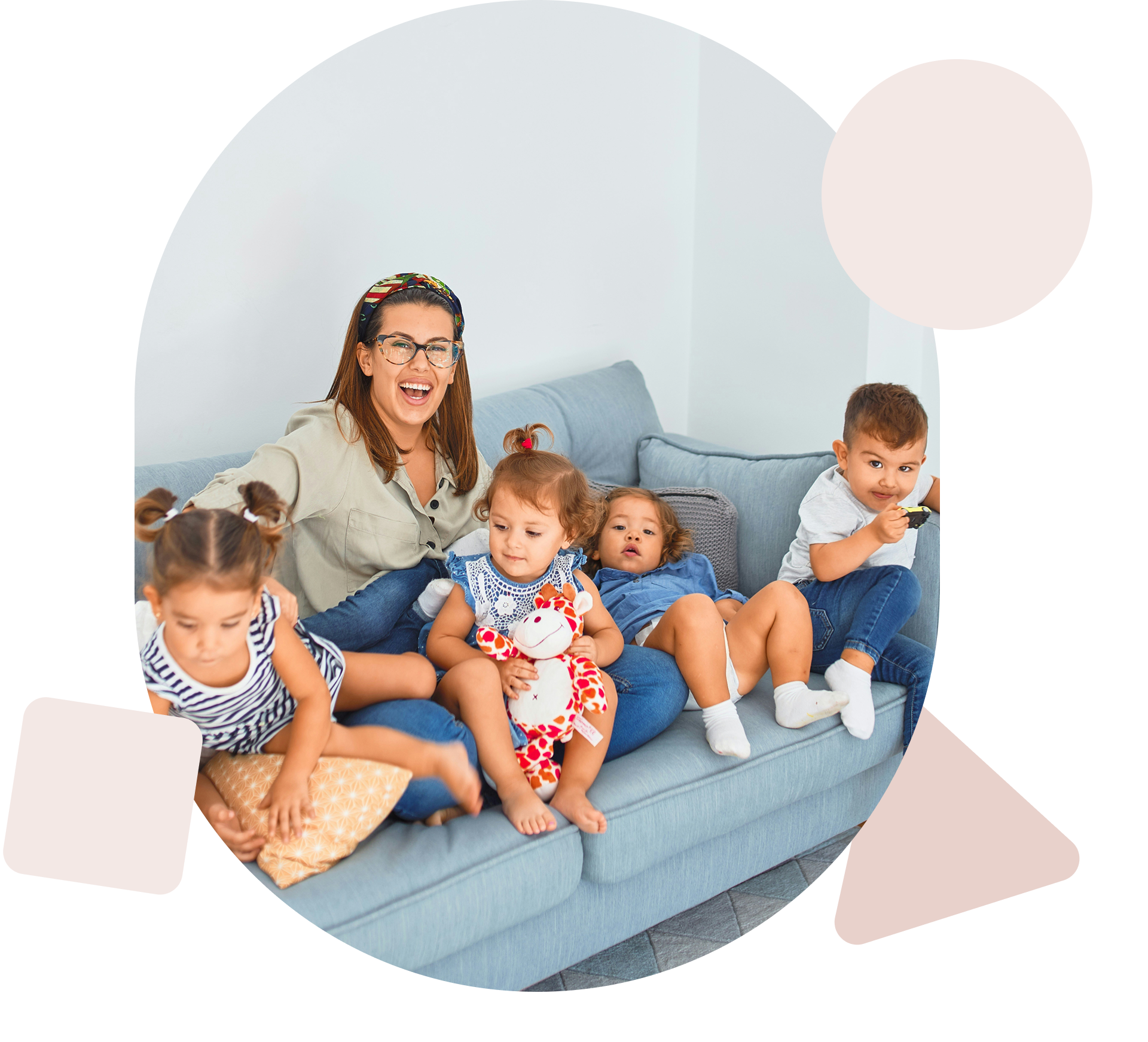 A woman with glasses and a colorful headscarf sitting on a blue sofa surrounded by young children, some holding toys and plush animals, in a bright room with white walls.