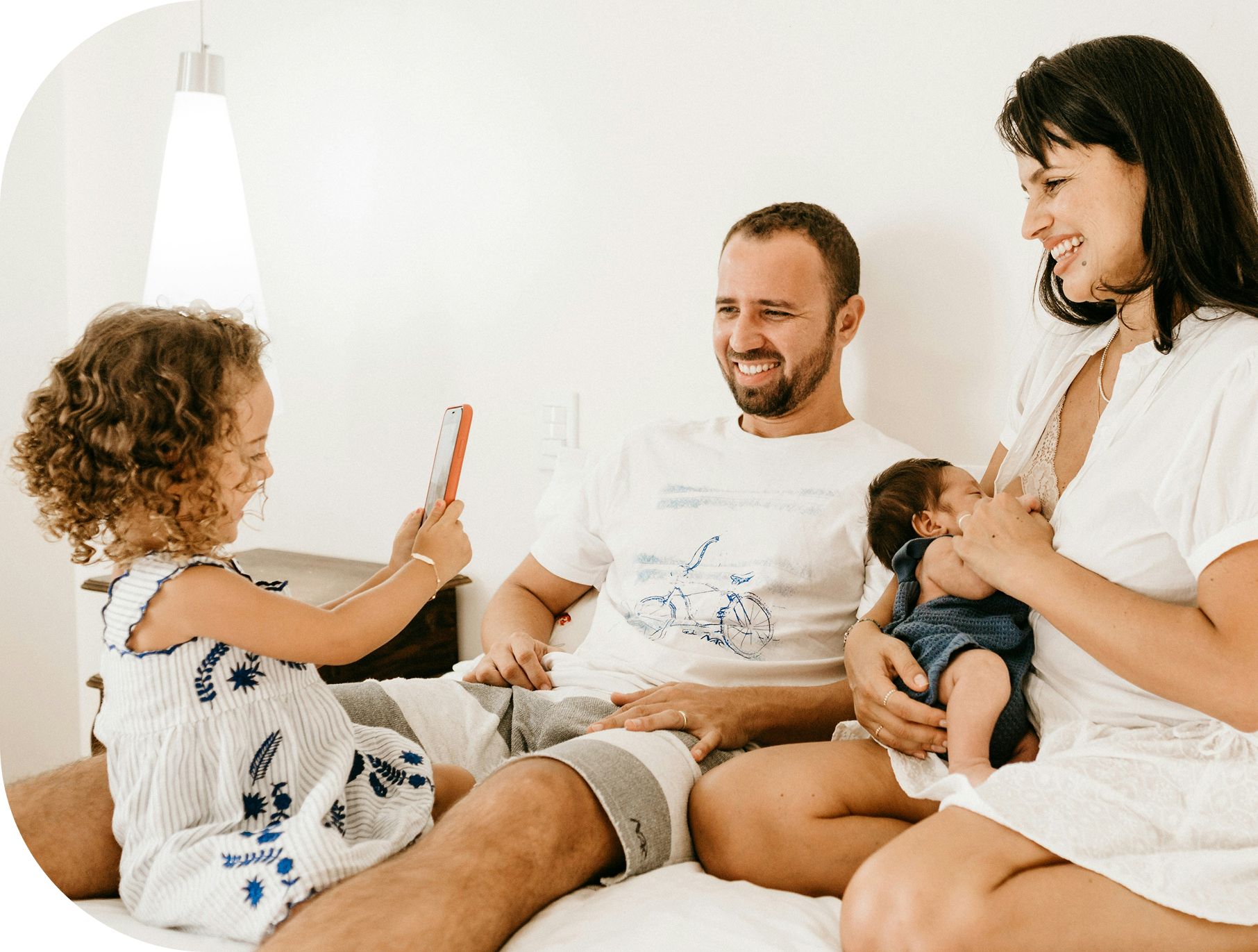 A family of five, including a girl, a man, a woman, and a baby, sitting on a bed in a bright room. The girl is taking a photo of the parents and baby with her phone. The woman is holding the baby while smiling, and the man is sitting next to them, also smiling.