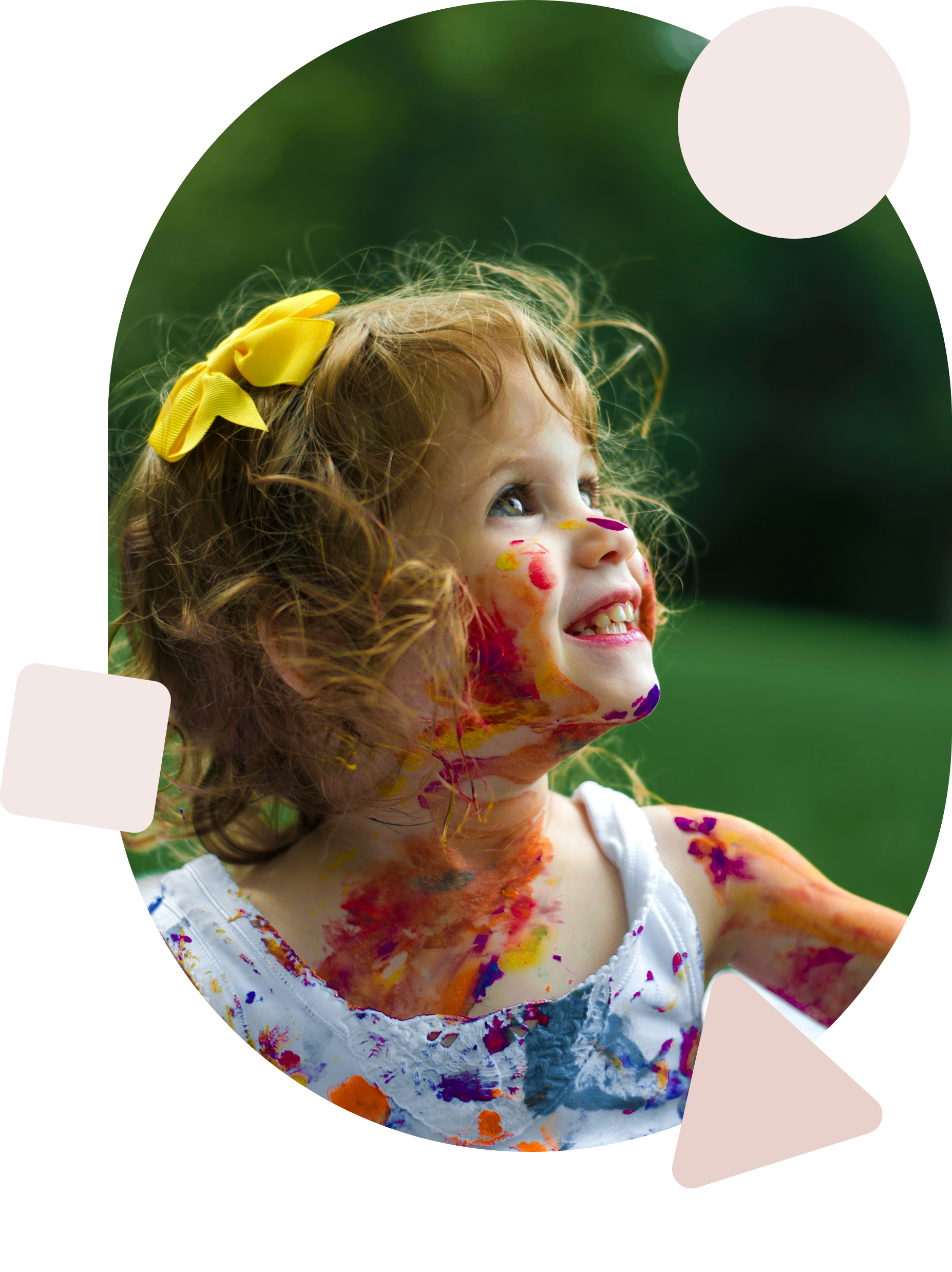 A young girl with curly red hair and a yellow bow, smiling outdoors, with colorful paint on her face and body, wearing a white sleeveless dress with embroidered details.
