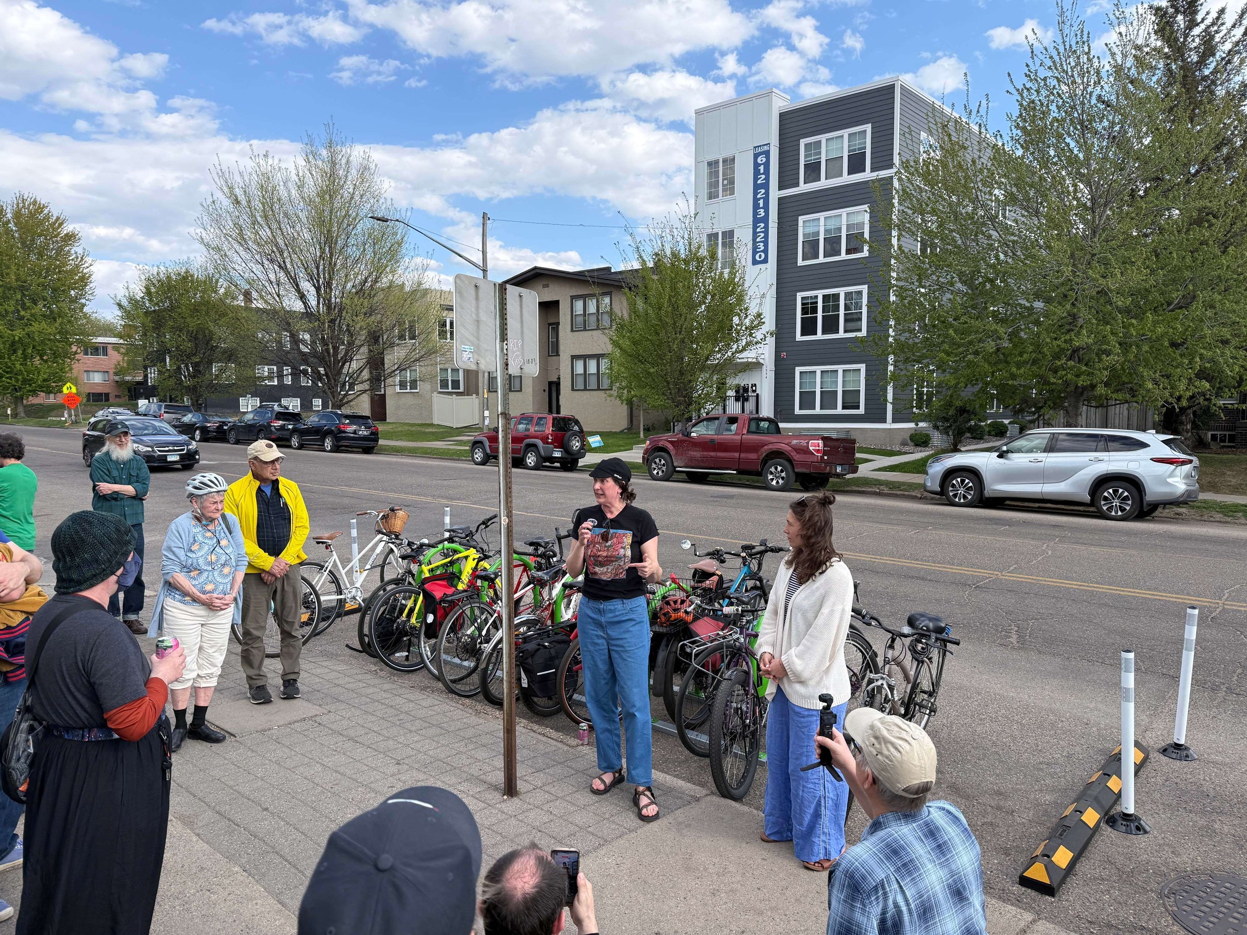 A group of people gathered on a sidewalk next to a full bike corral, with some speaking and others listening. Behind them are several parked cars and residential buildings, with trees and a partly cloudy sky above.