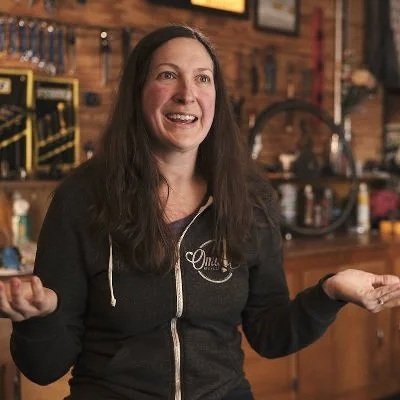 A woman smiling with arms out, palms up, as if asking a question, in a bike shop setting with a full tool board behind her.