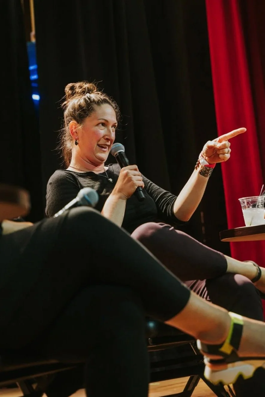 A woman sitting on stage, smiling and holding a microphone, pointing with her left hand. She is wearing a black top, dark pants, and black sandals. Red and black curtains are in the background because she's on a stage.