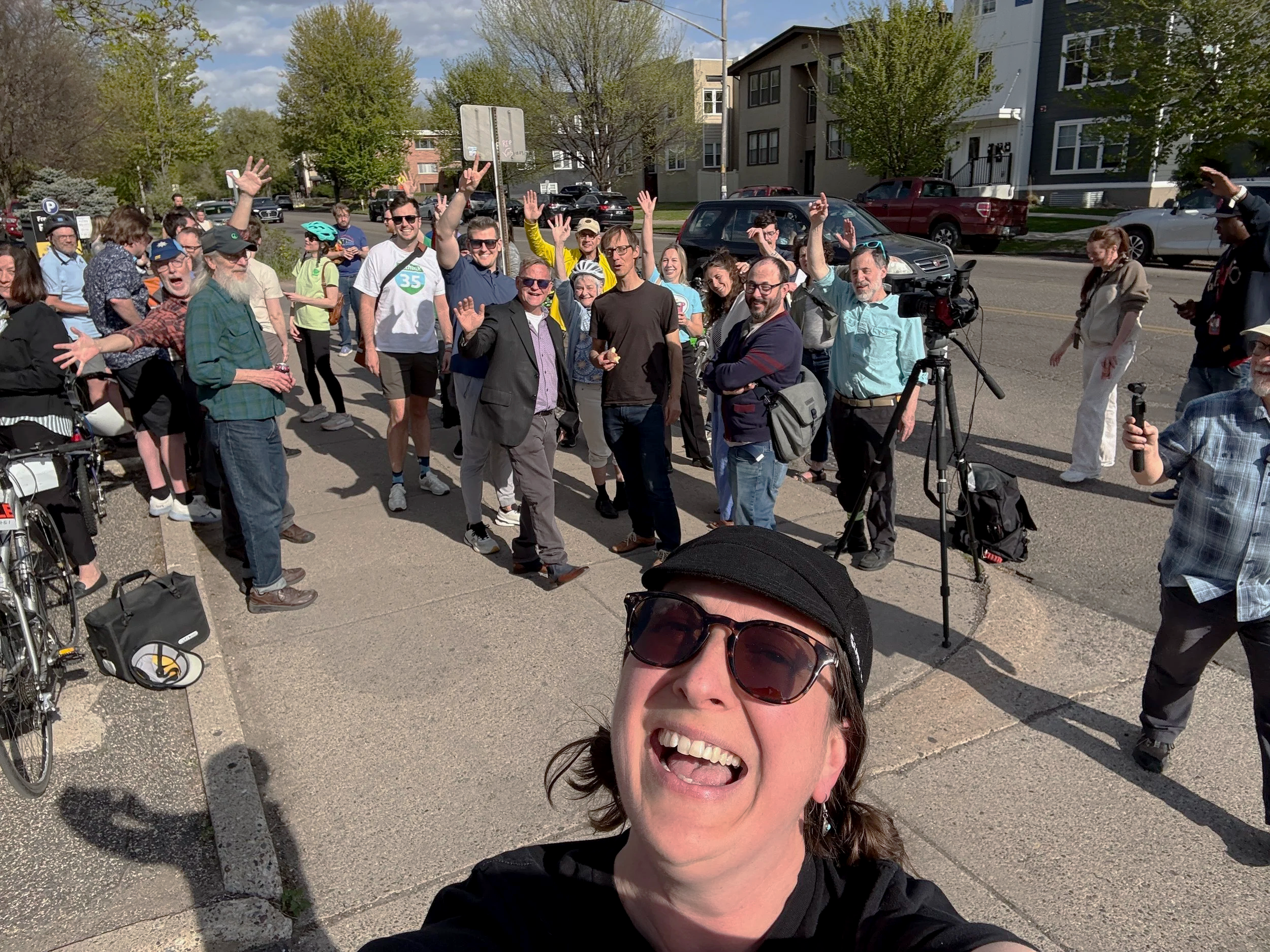 Selfie of a woman with sunglasses smiling in the foreground with a crowd of people in the background by a new bike corral, outdoors on a sunny day with buildings and trees.