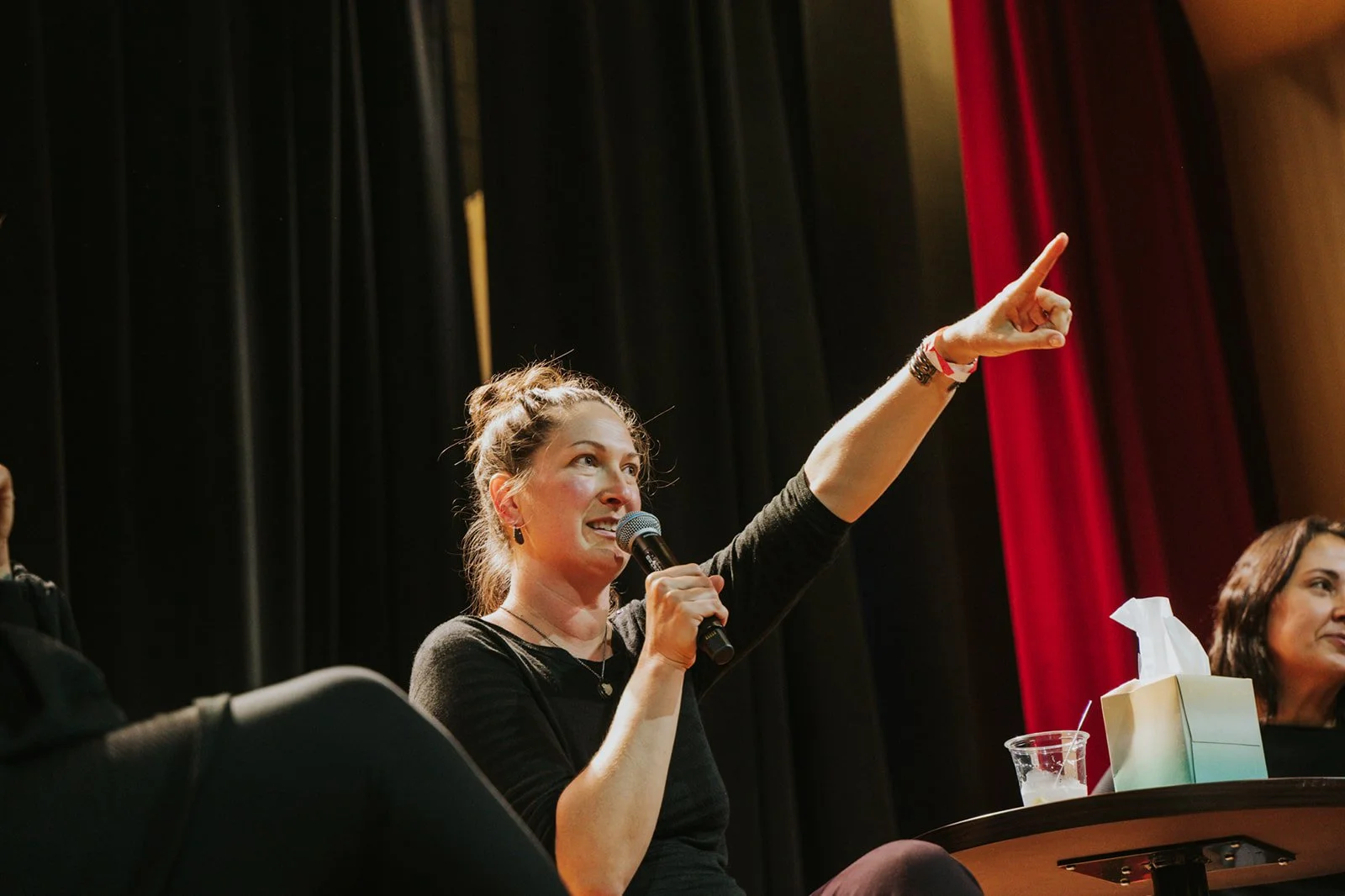 A woman with brown hair in a bun, wearing a black shirt, speaking into a microphone and gesturing with her left hand while sitting at a panel. There is another woman with dark hair sitting next to her, a tissue box, and a plastic cup on the table.