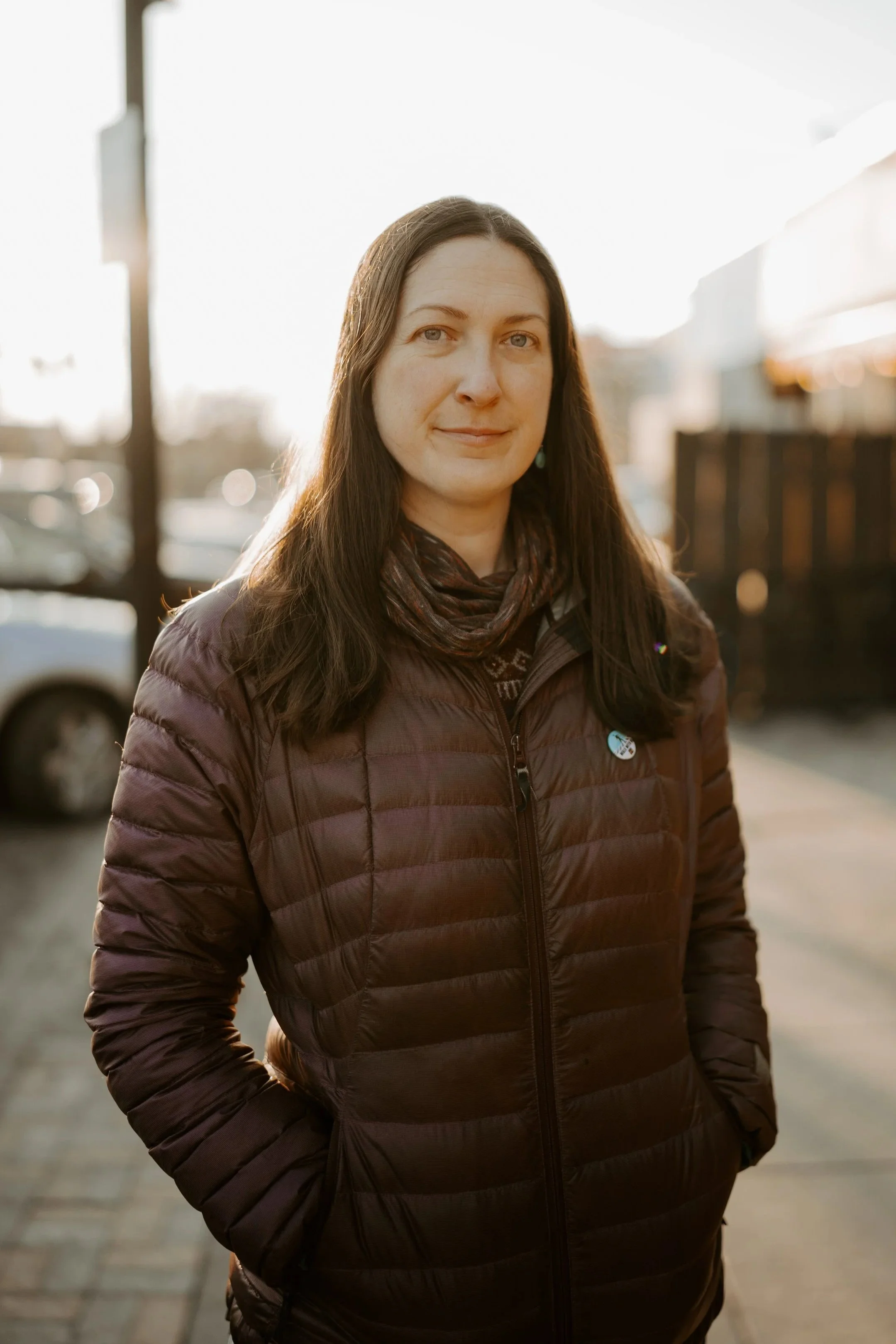 A woman with long brown hair and blue eyes standing outdoors in sunlight, wearing a dark purple jacket and a patterned scarf.