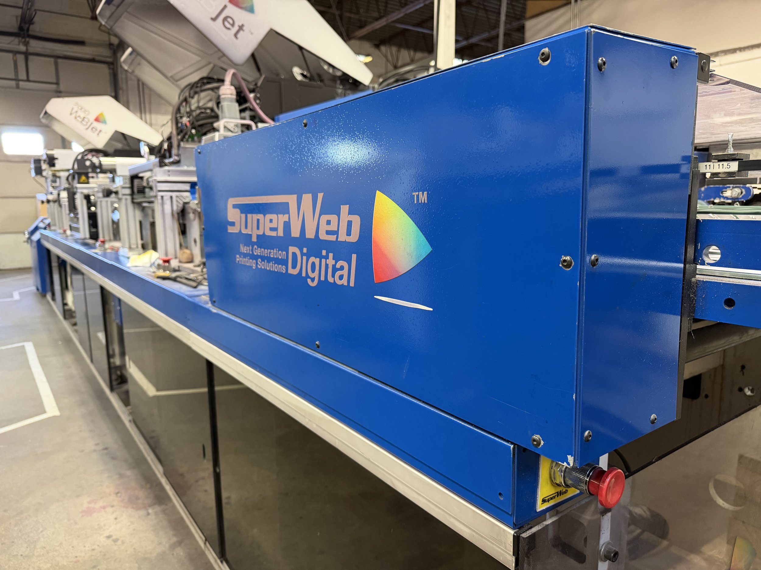 Blue industrial machine labeled 'SuperWeb Digital' with a rainbow-colored logo, situated on a metal frame in an indoor industrial setting.