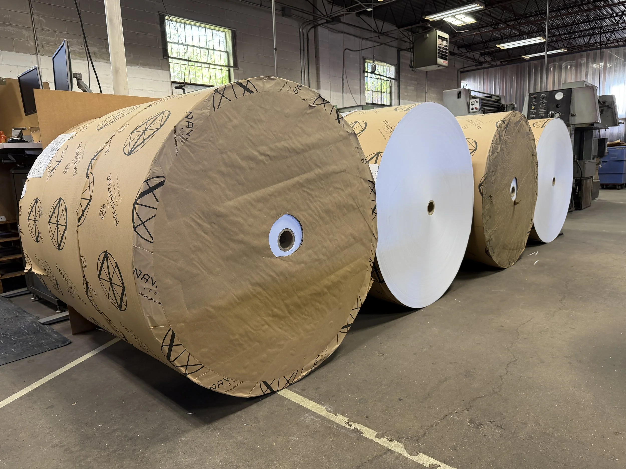 Large industrial paper rolls in a warehouse with high ceiling, fluorescent lighting, and small windows, next to machinery.
