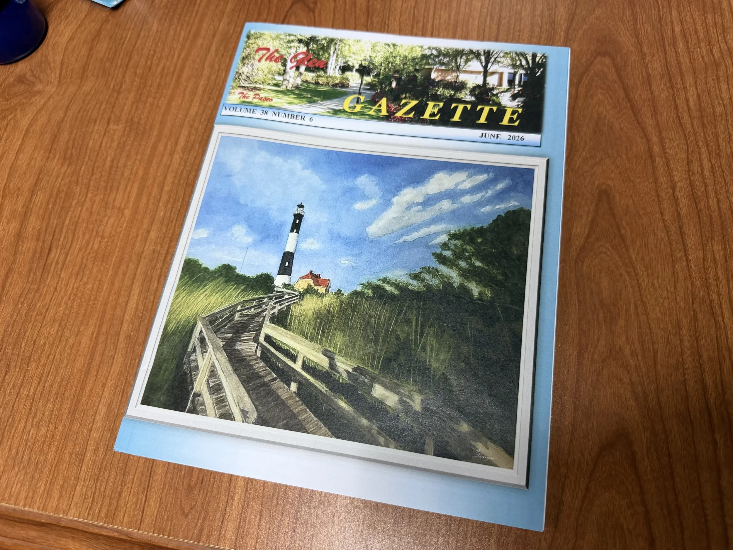 A magazine lying on a wooden table, July 2026 issue. The cover features a painting of a lighthouse with a cloudy sky, a path, and trees.