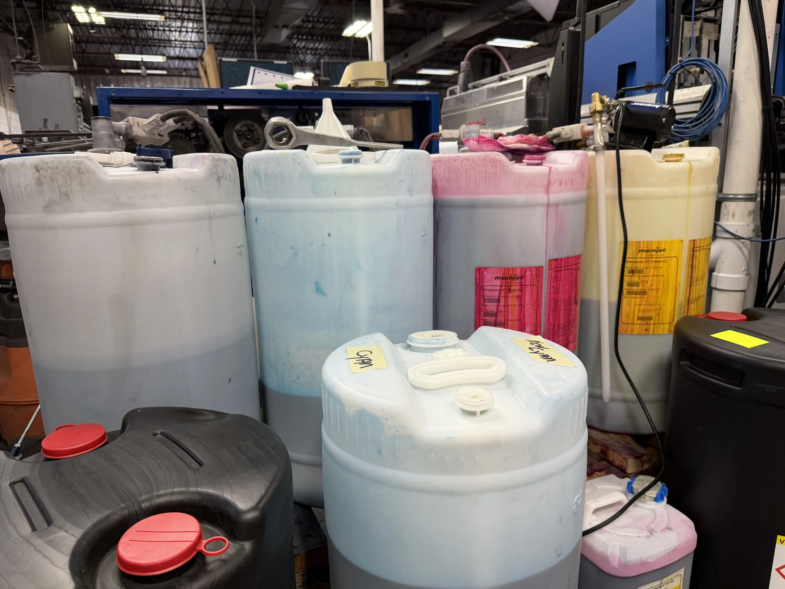 Multiple plastic containers of various colors and sizes stored on a workbench or shelf in an industrial or warehouse setting, with hoses and machinery in the background.