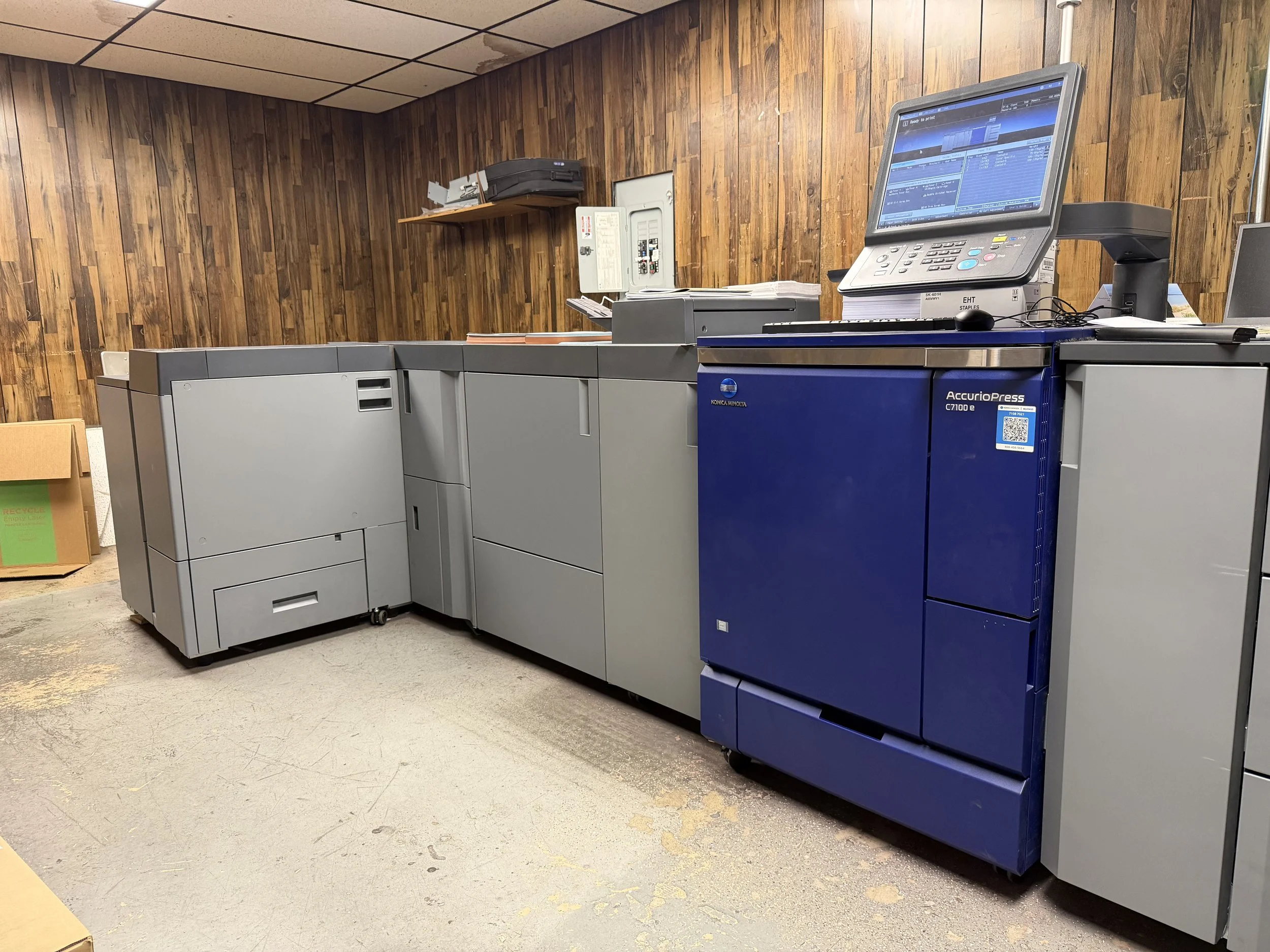 A blue and gray Konica Minolta AccurioPress digital printing press in an office or print shop next to a gray machine. There are papers, a computer monitor, and office supplies on top, with wood panel walls in the background.