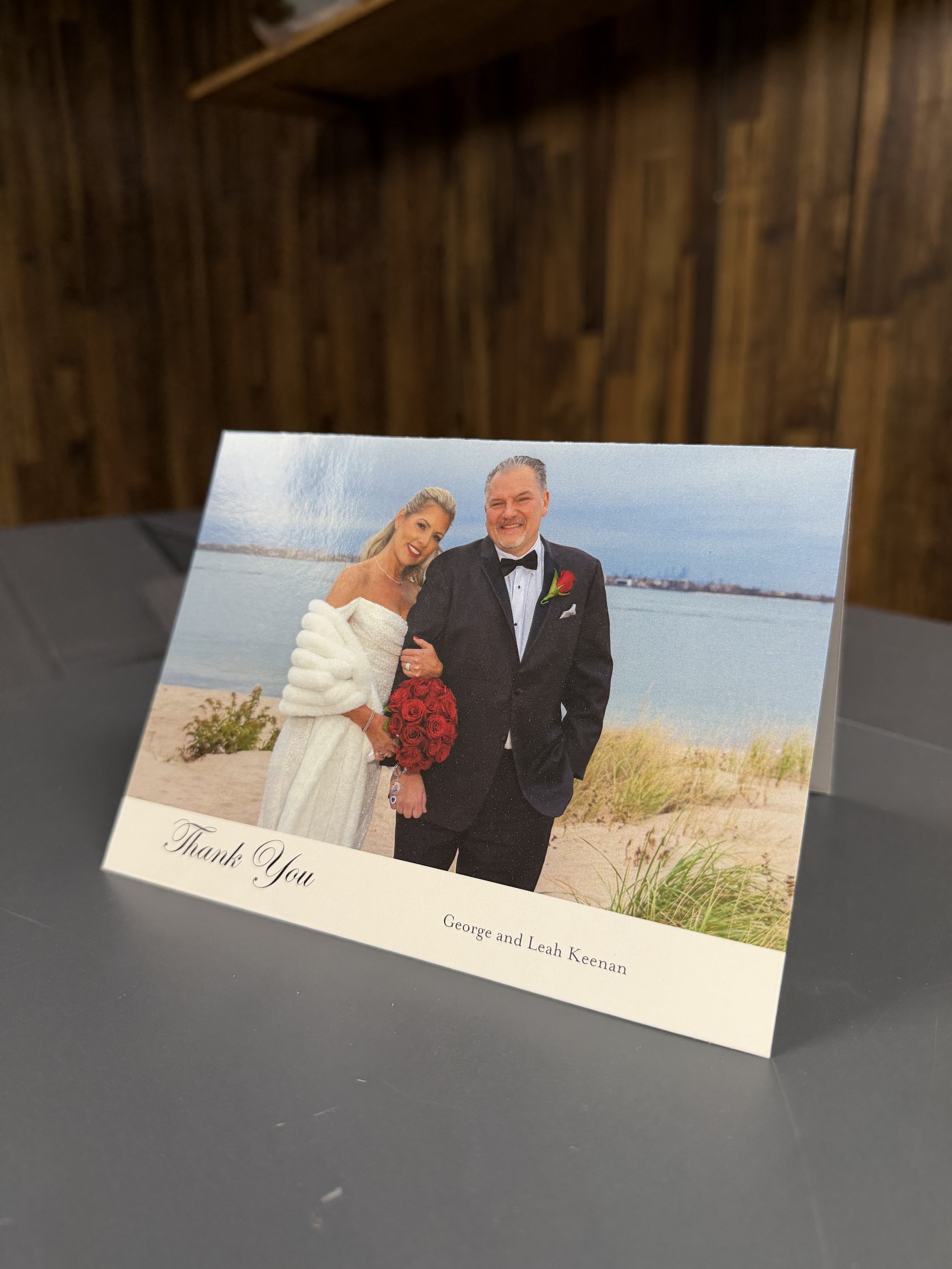 A wedding thank you card featuring a photo of a bride and groom on a beach, with the bride in a white dress holding a bouquet of red roses, and the groom in a black tuxedo. The card has a handwritten style message that reads "Thank You"