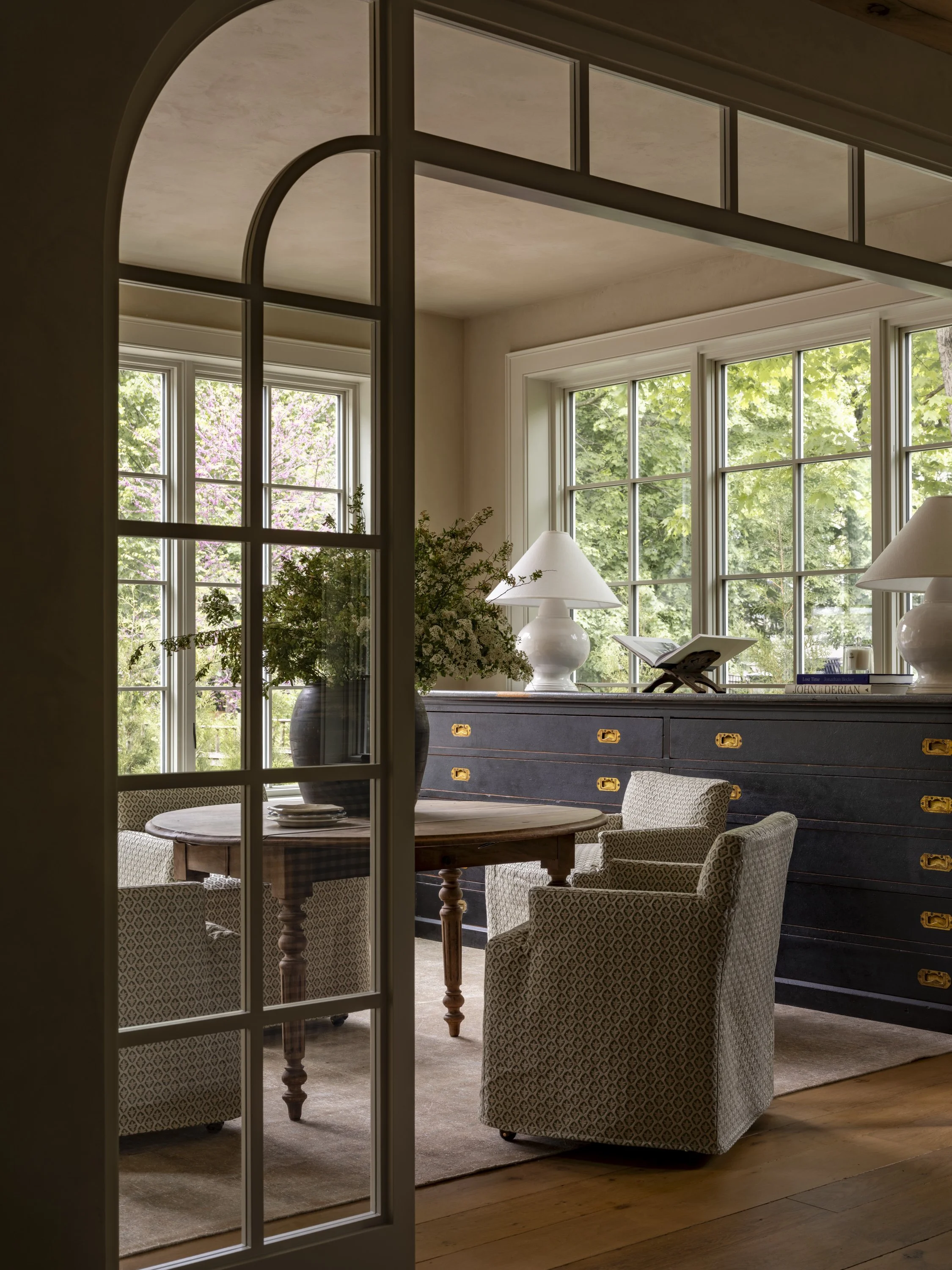 Home study in a Cottagewood residence viewed through arched steel and glass doors with custom cabinetry, oversized windows, and warm neutral furnishings.