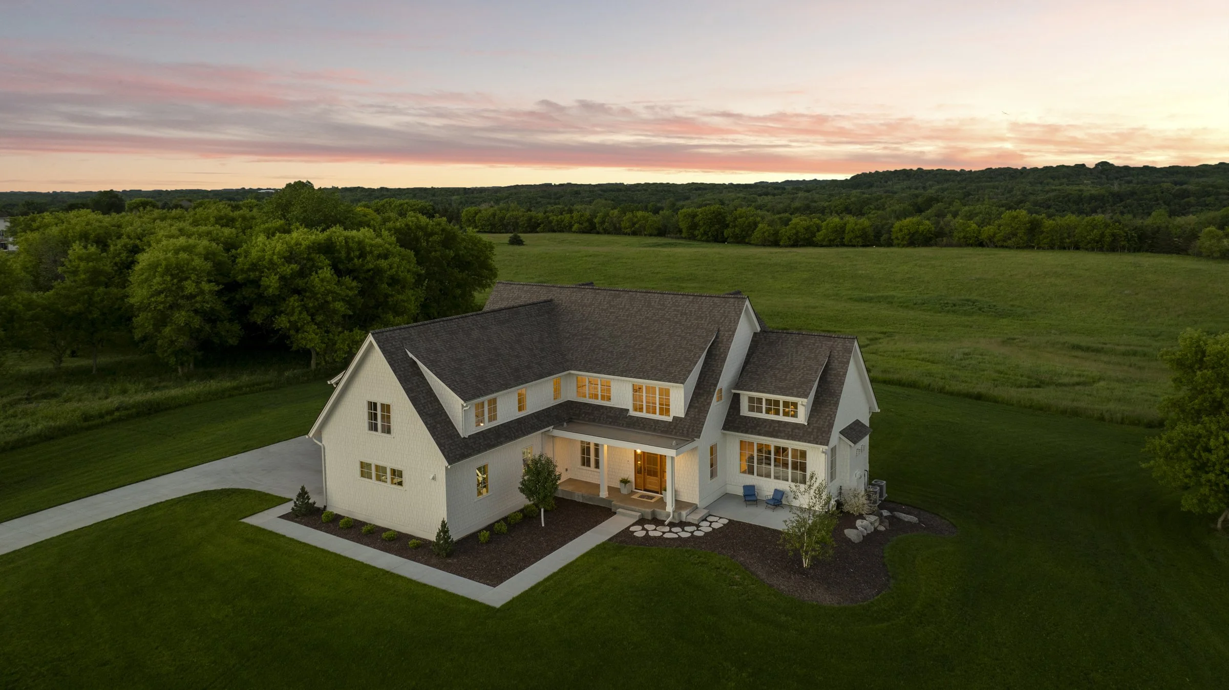 Aerial view of a custom home in Rochester, Minnesota surrounded by rolling countryside and mature trees at sunset, showcasing timeless architecture and expansive rural views.