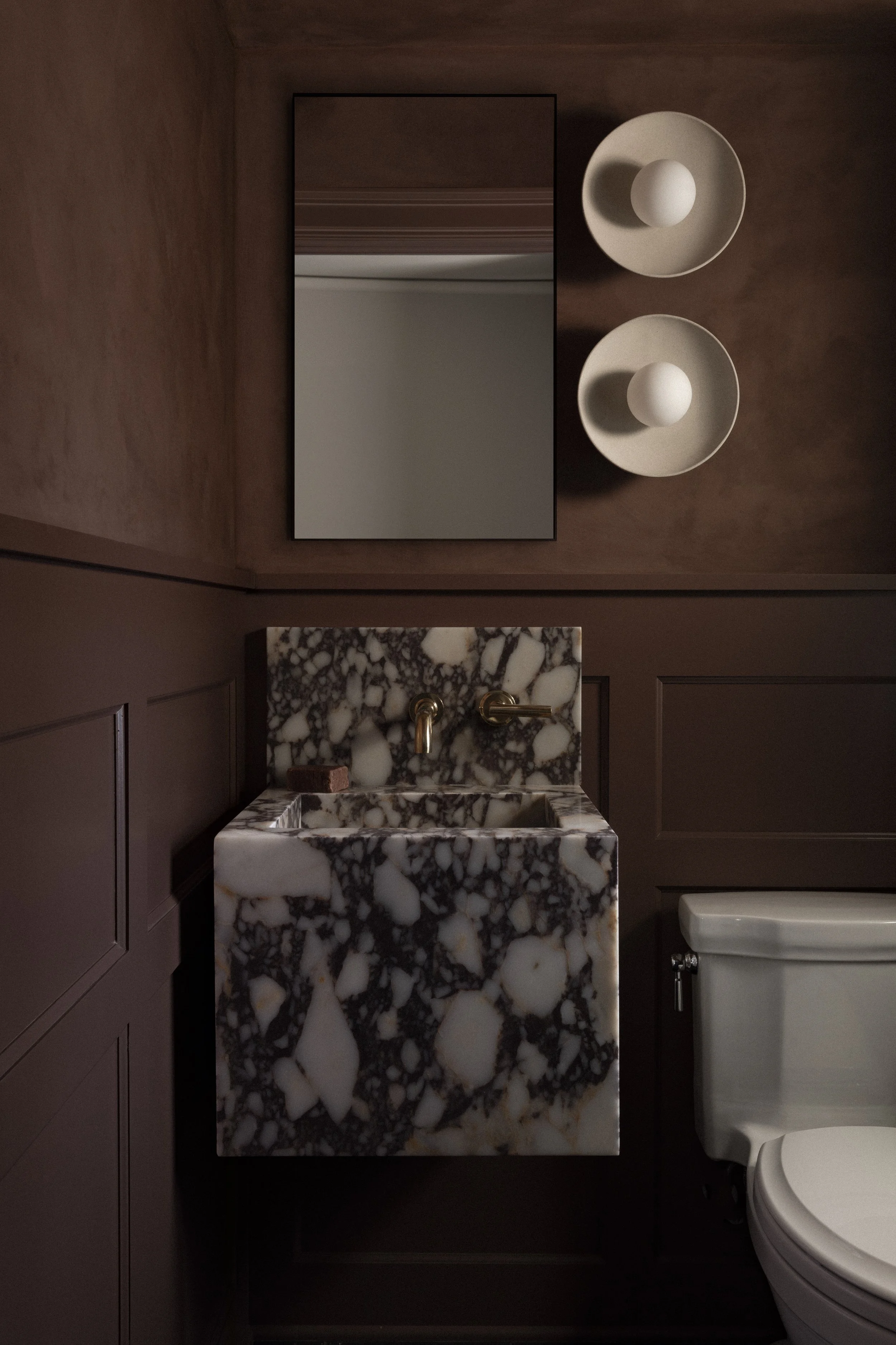 Powder bath in a Minneapolis remodel featuring a sculptural marble sink, moody wall color, and brass wall-mounted fixtures.