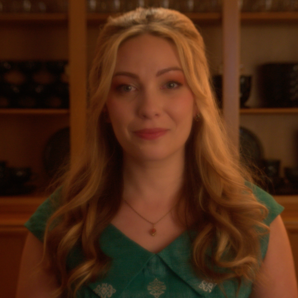 A woman with long, wavy blonde hair smiling in front of wooden shelves filled with pottery.