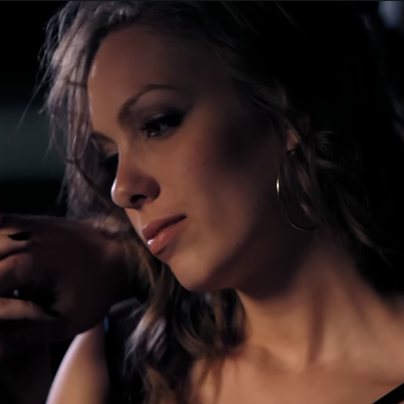 Close-up of a woman with wavy hair, wearing hoop earrings, looking down thoughtfully in a dimly lit setting.