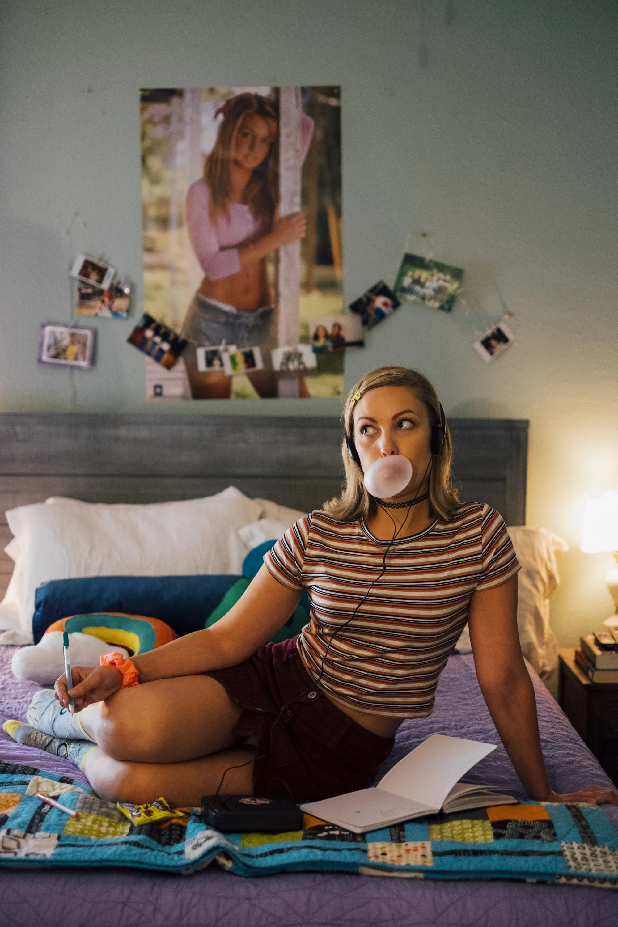 A young woman sits on a bed wearing a striped shirt and maroon shorts, blowing a bubble gum bubble, with headphones around her neck, a notebook on her lap, and a pen in her hand. Behind her, there's a colorful quilt, and photographs hanging on the wa