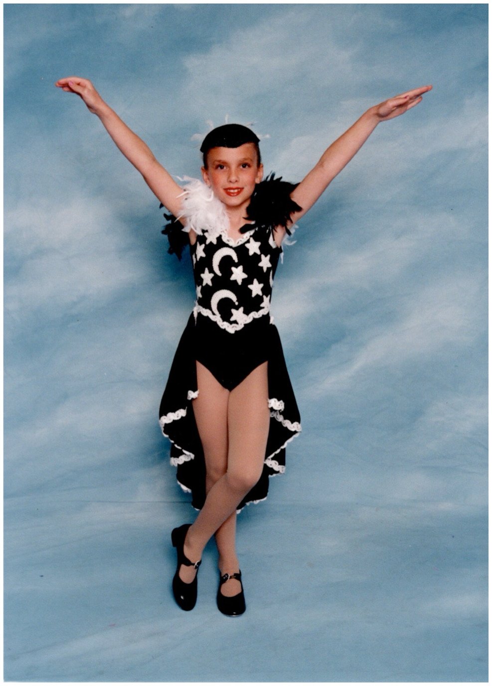 A young girl in a black and white costume with star and moon designs, feather accents, and a skirt reveals her legs, posing with arms raised and smiling against a blue sky backdrop.