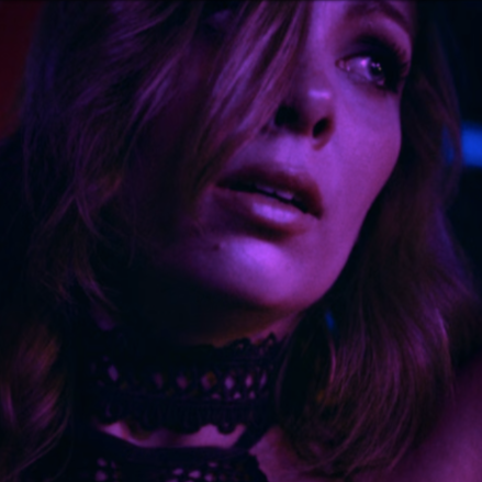 Close-up of a woman with wavy hair and makeup, illuminated by purple light.