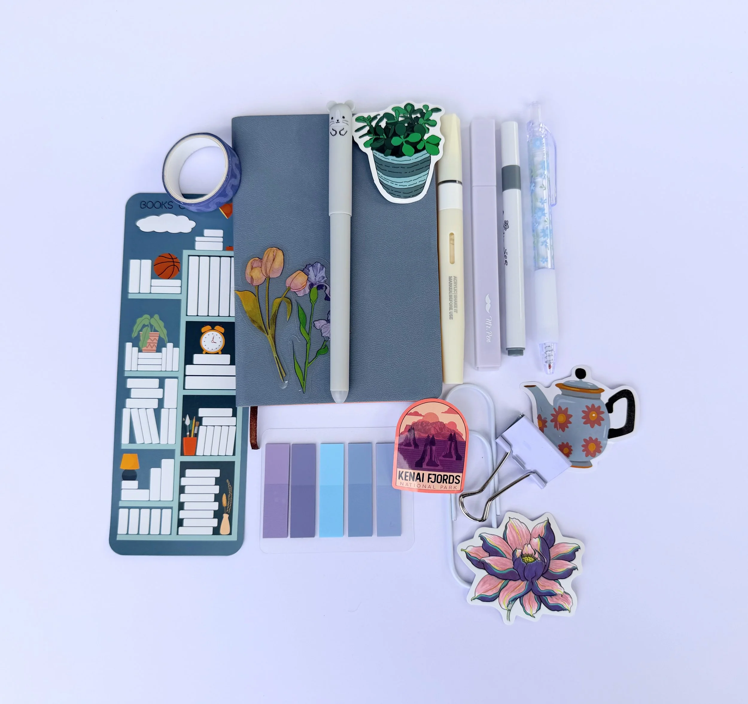 Flat lay of stationeries including notebooks, pens, stickers, washi tape, a paperclip, and decorative items on a white background.