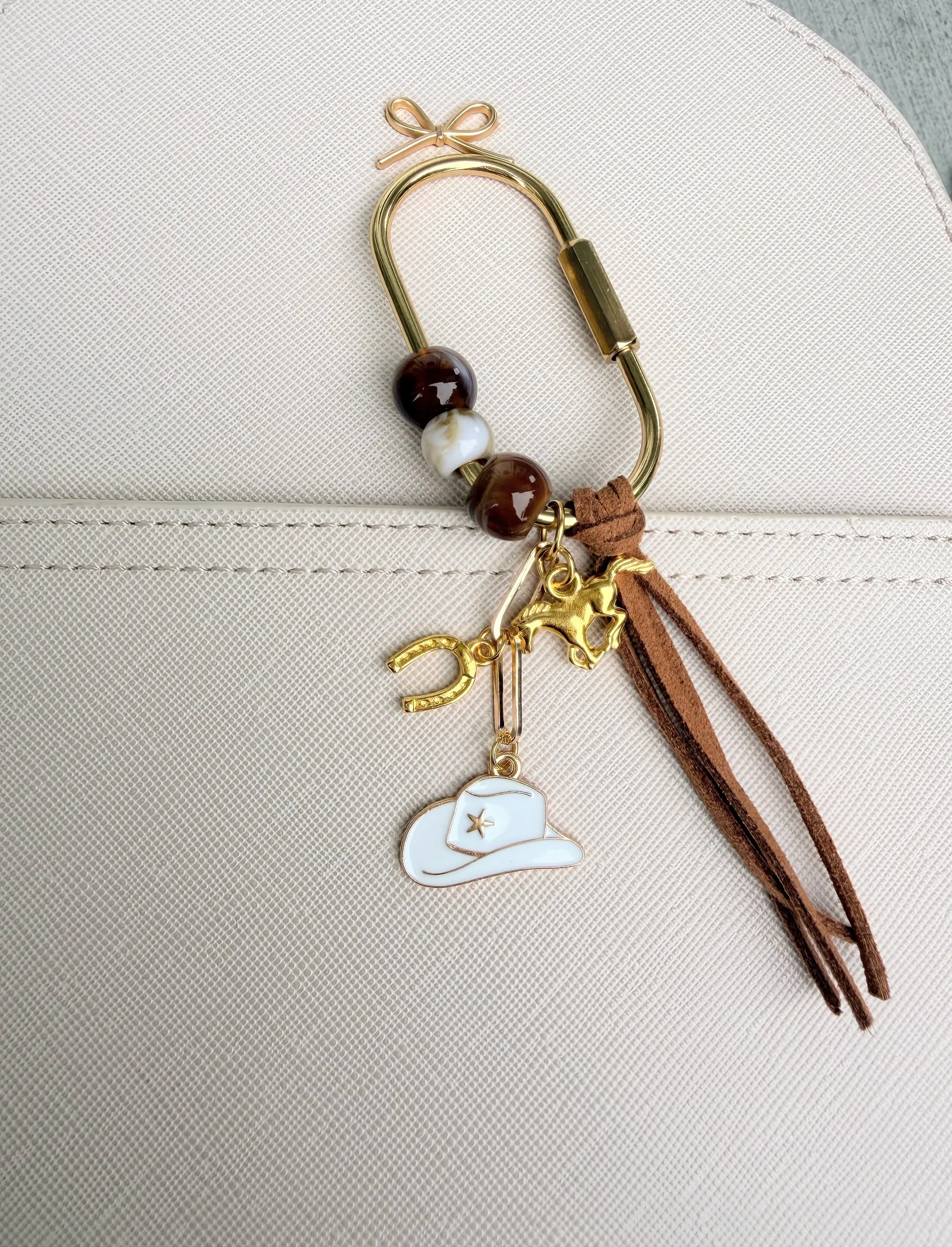 A close-up of a beige textured surface with a decorative metallic bookmark with gold and pink accents. The bookmark includes a charm in the shape of a hat with a star, and there are brown leather and suede strings attached.